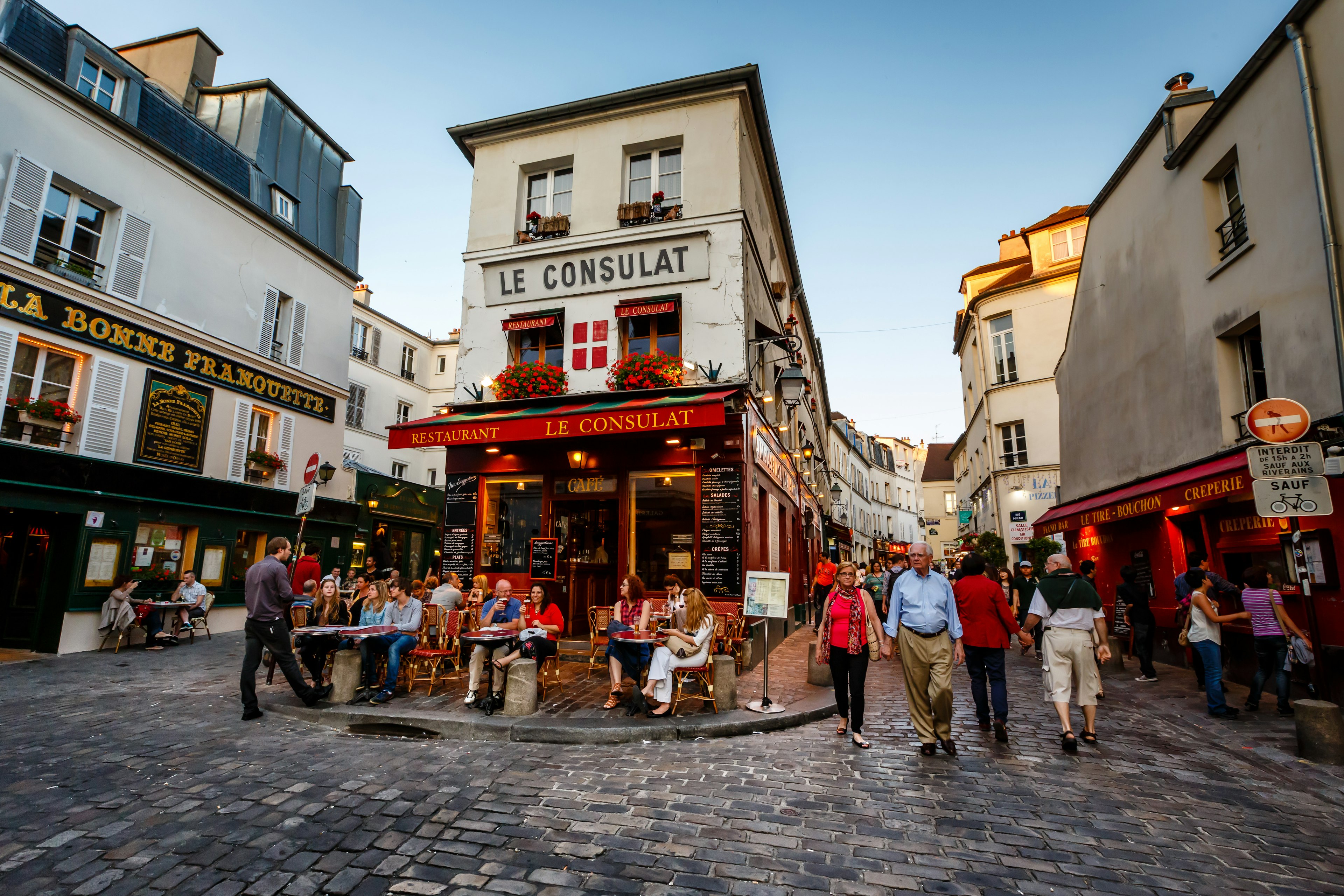 People sit at tables outside Le Consulat, a popular cafe in Montmartre, Paris. Other people walk along the pretty cobbled streets either side of the cafe.