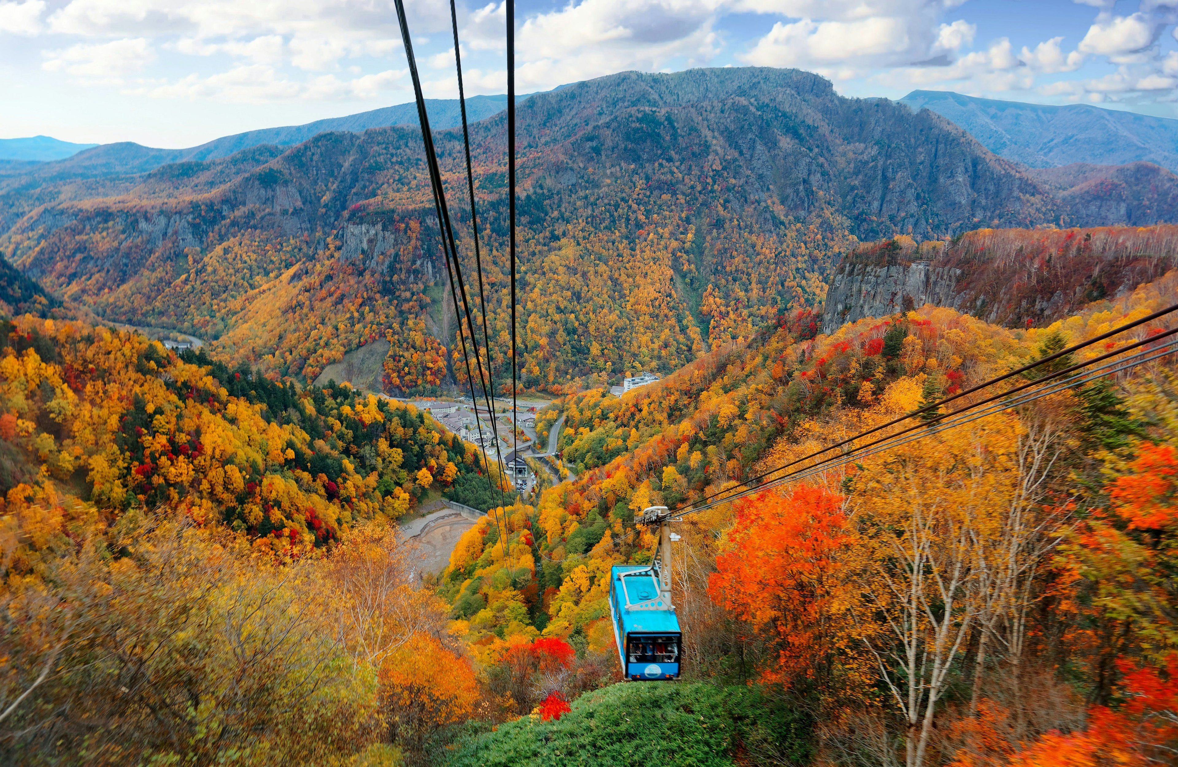 A blue cable car carriage travels up through trees changing from green to orange, red and yellow during fall