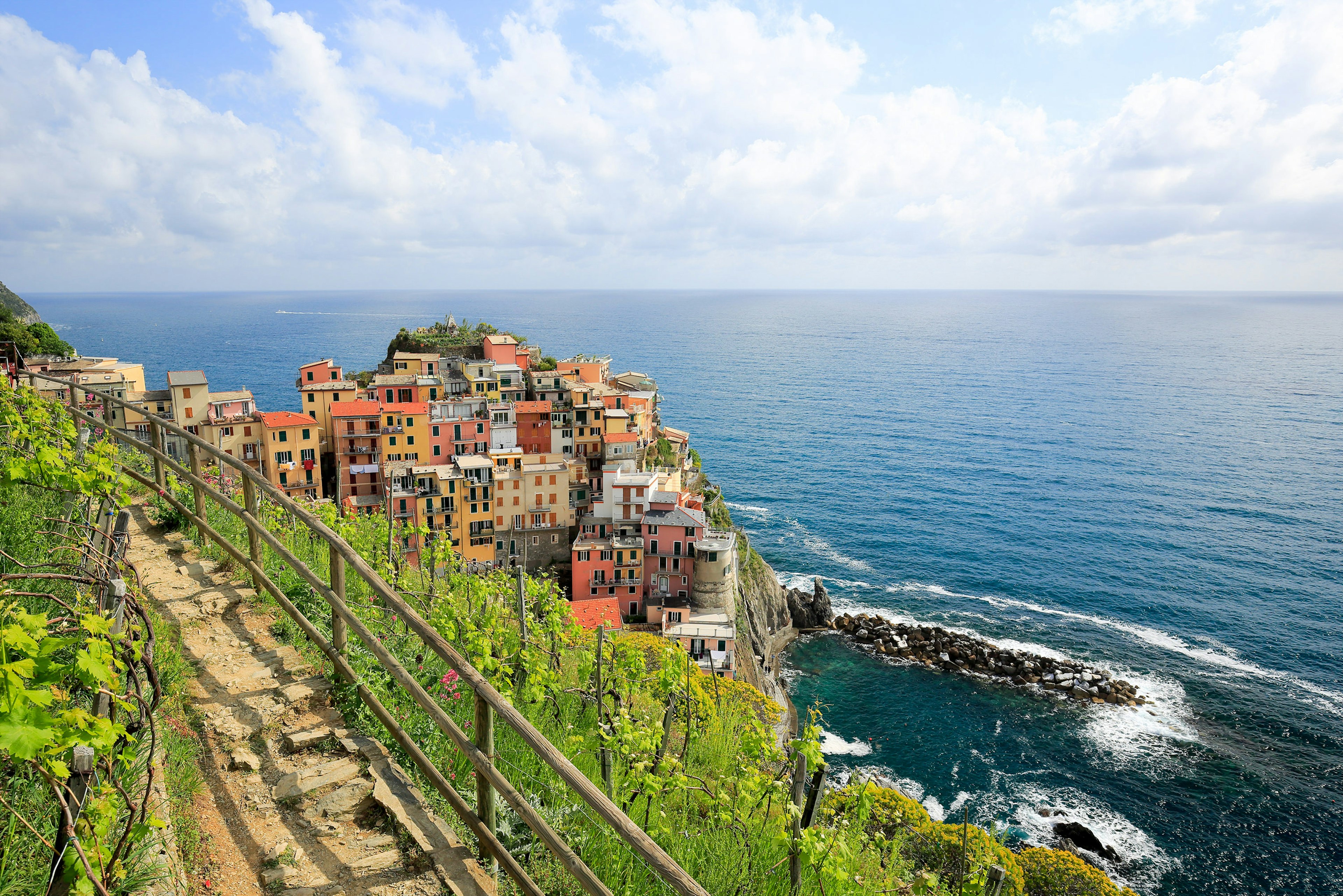 A walking path along the coast surrounded by vineyards with a colorful village built onto a rocky outcrop at sea