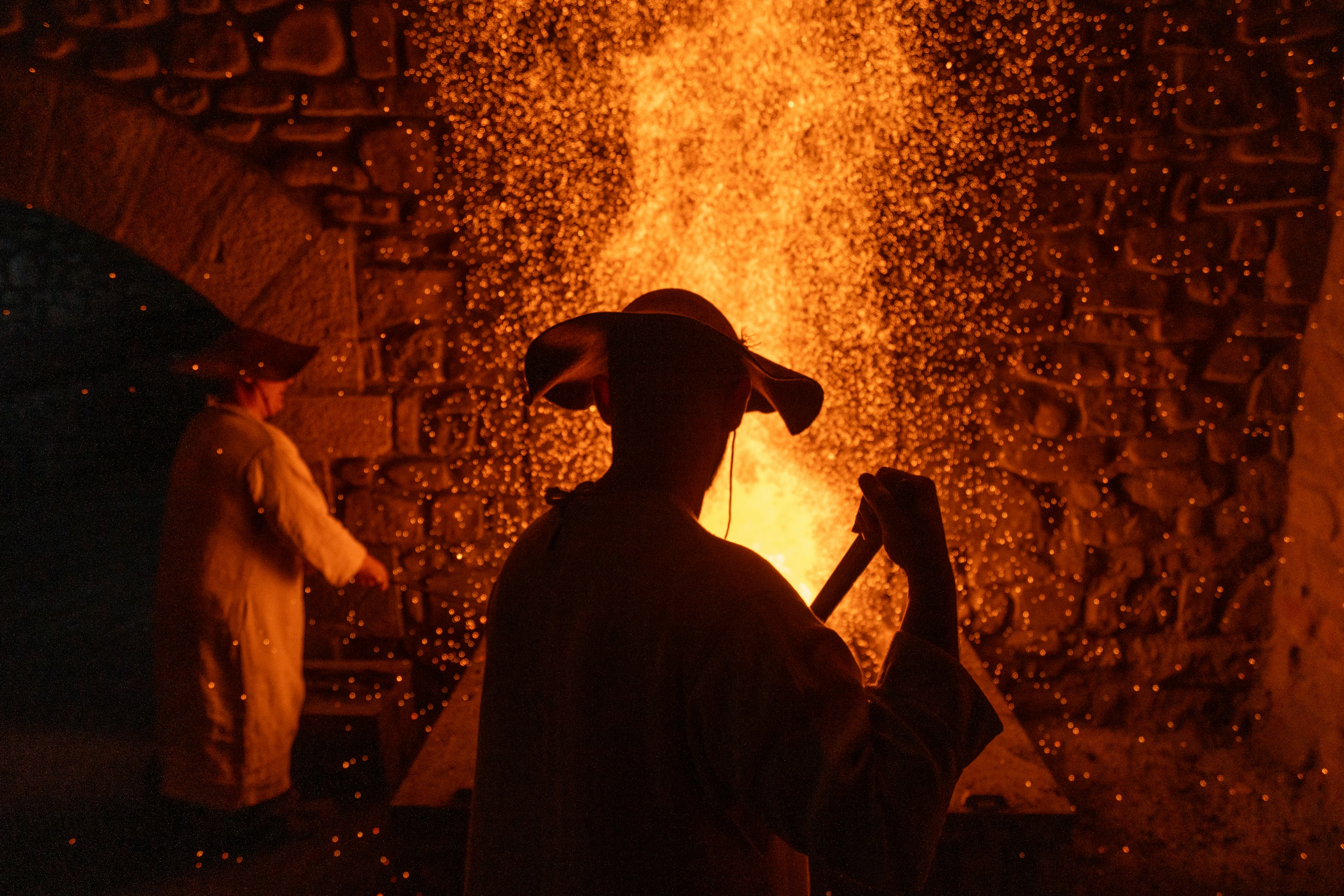 An ironworker holding a tool is silhouetted against flames and sparks at a foundry