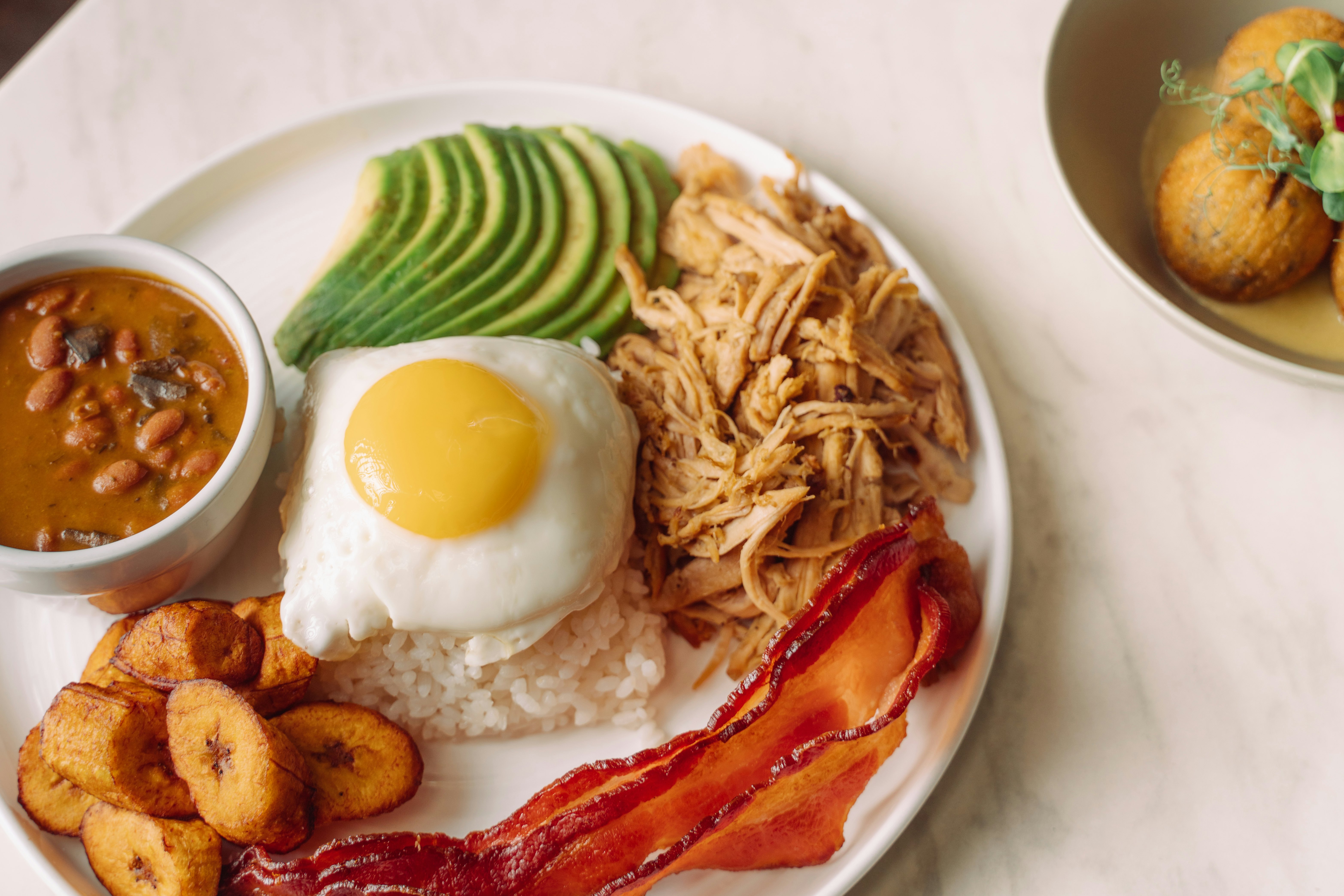 A large Puerto Rican breakfast plate with plantains, bacon, egg, avocado, rice, beans and perníl