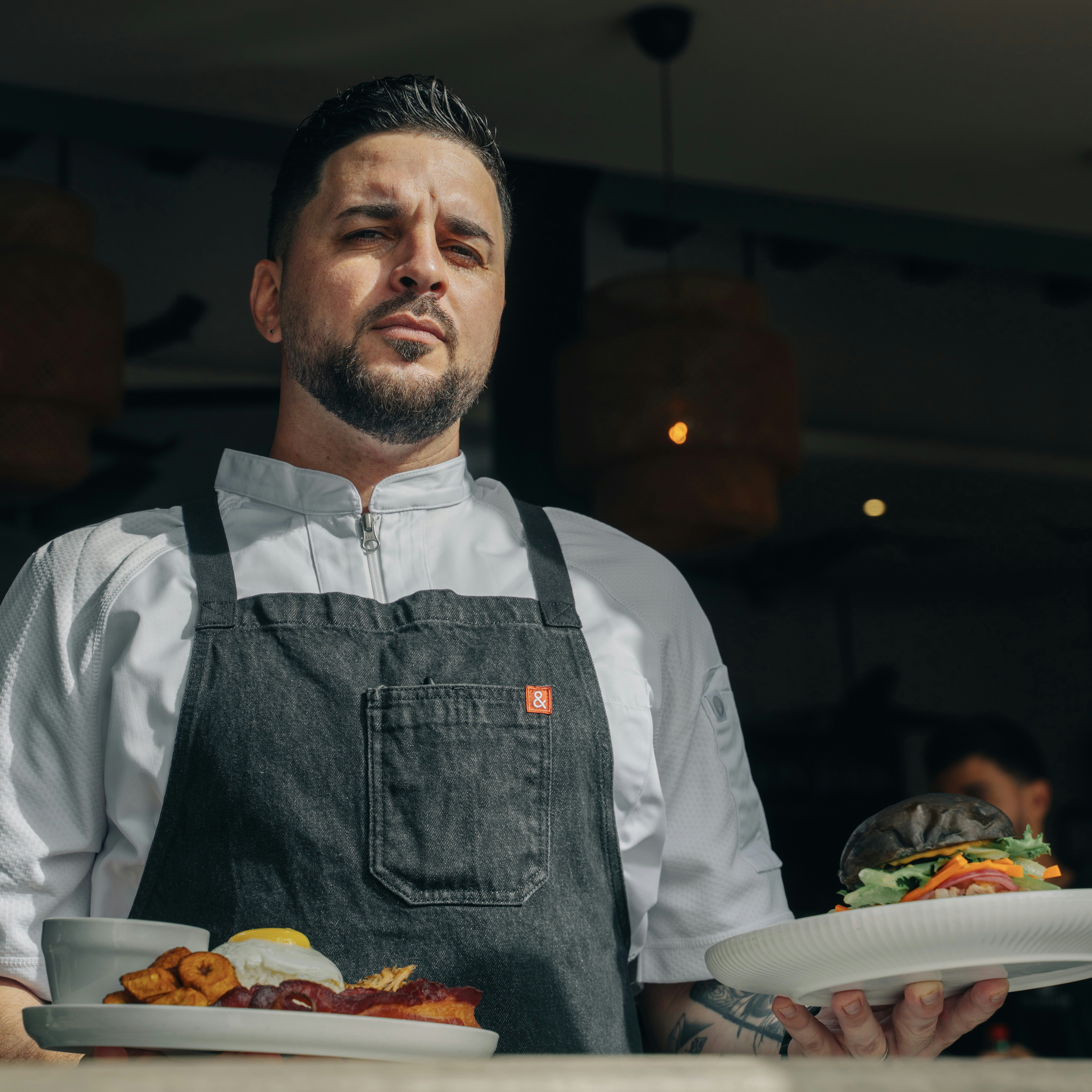 A chef with a neat beard in a denim apron and white uniform holds two plates of food