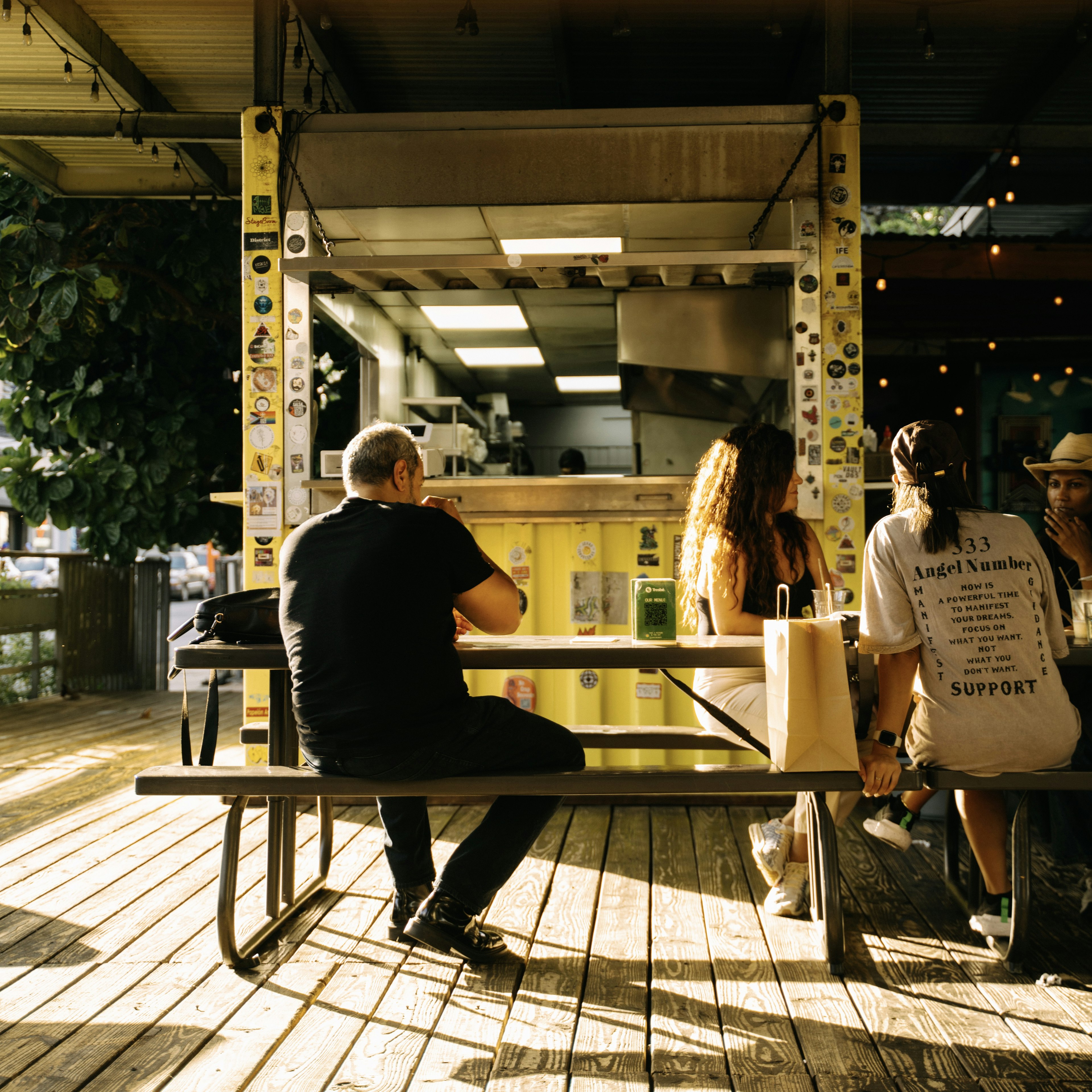 A group of people enjoy food at a picnic table on a deck next to a restaurant that occupies a converted yellow shipping container