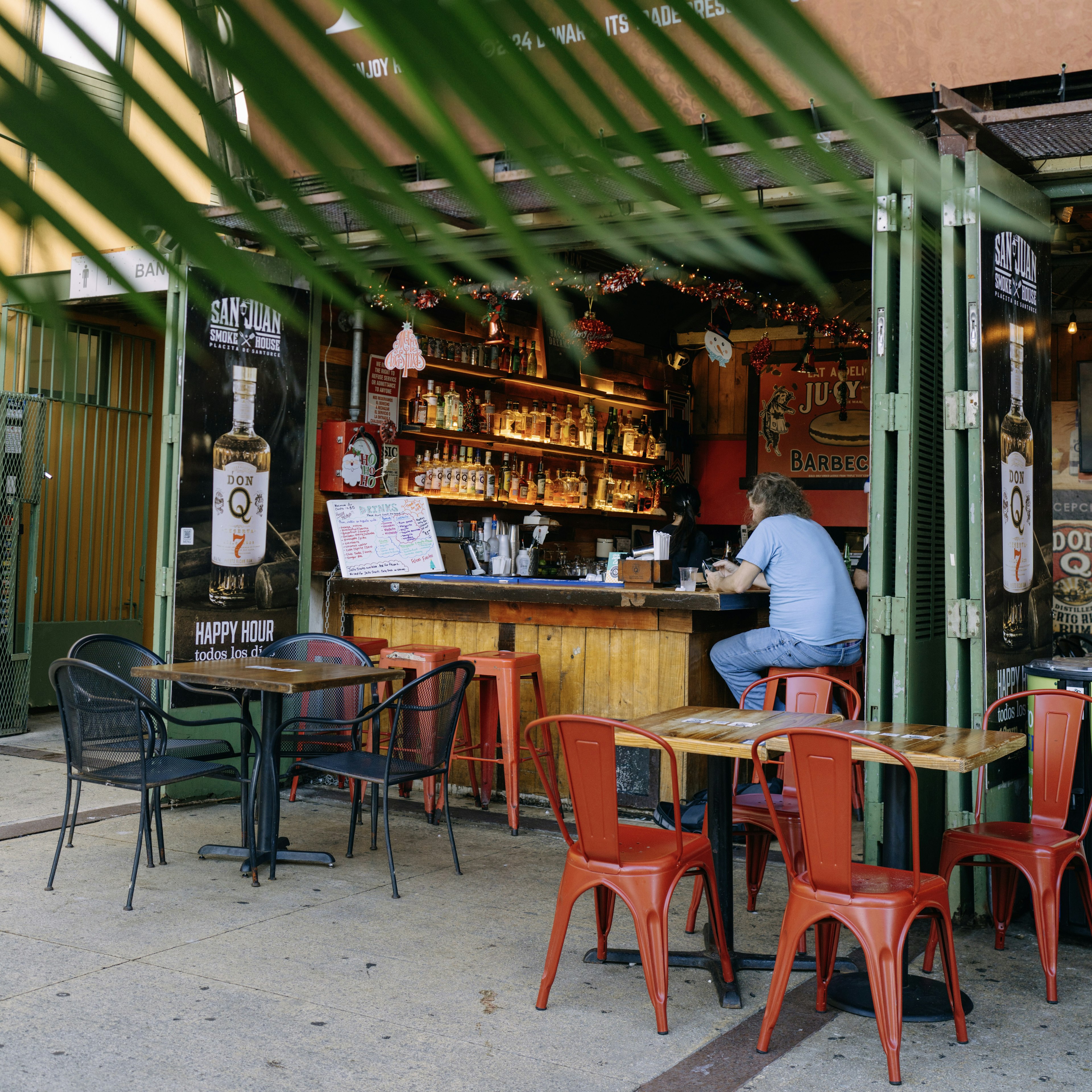 A man sits alone at a bar within a restaurant opening on to the street. Red and black chairs are set at sidewalk tables, while bottles on the bar wall are illuminated from beneath