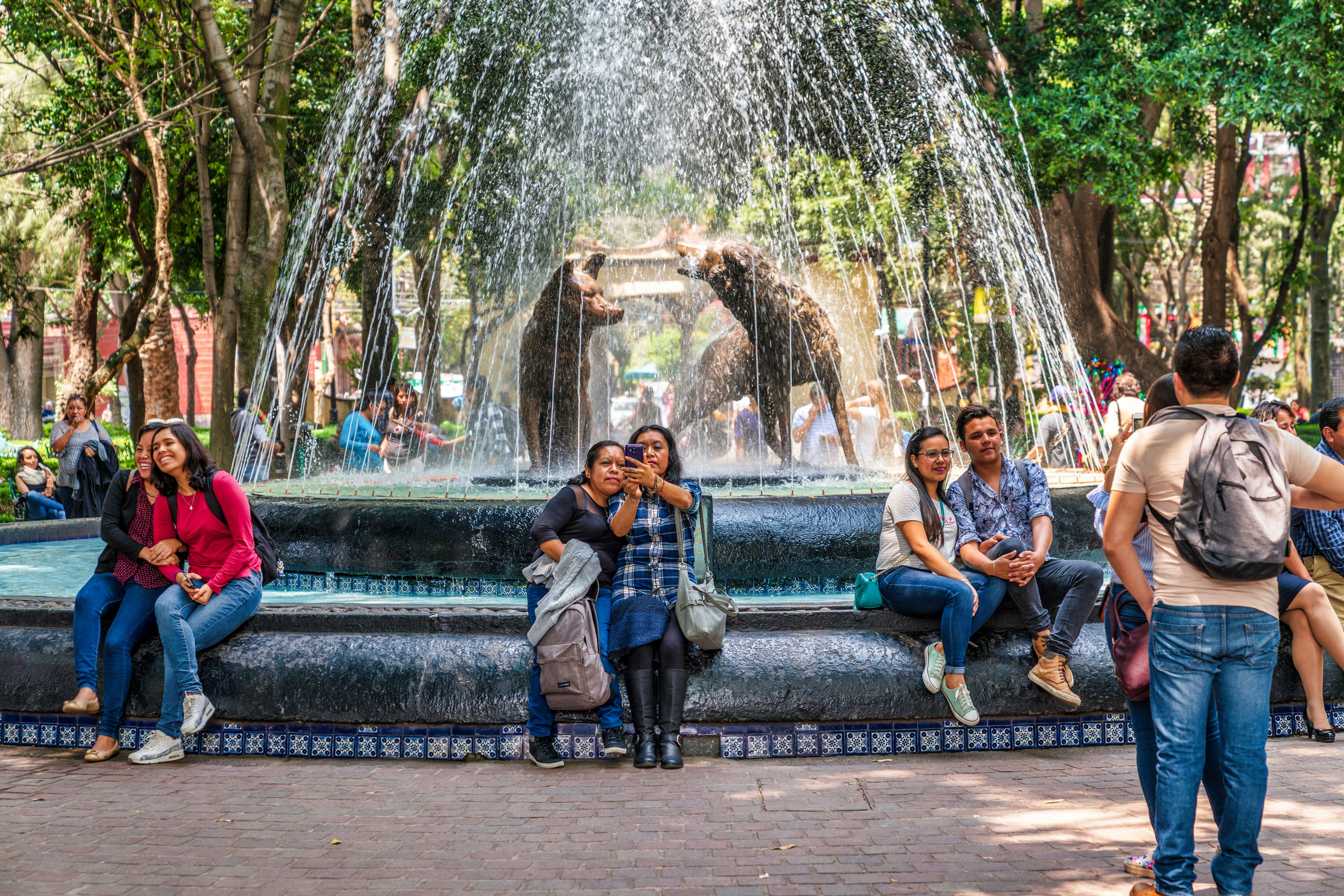 People sit and pose for pictures on the lip of a fountain with sculptures of wolves and high jets of water in a city park. Trees are visible in the background.