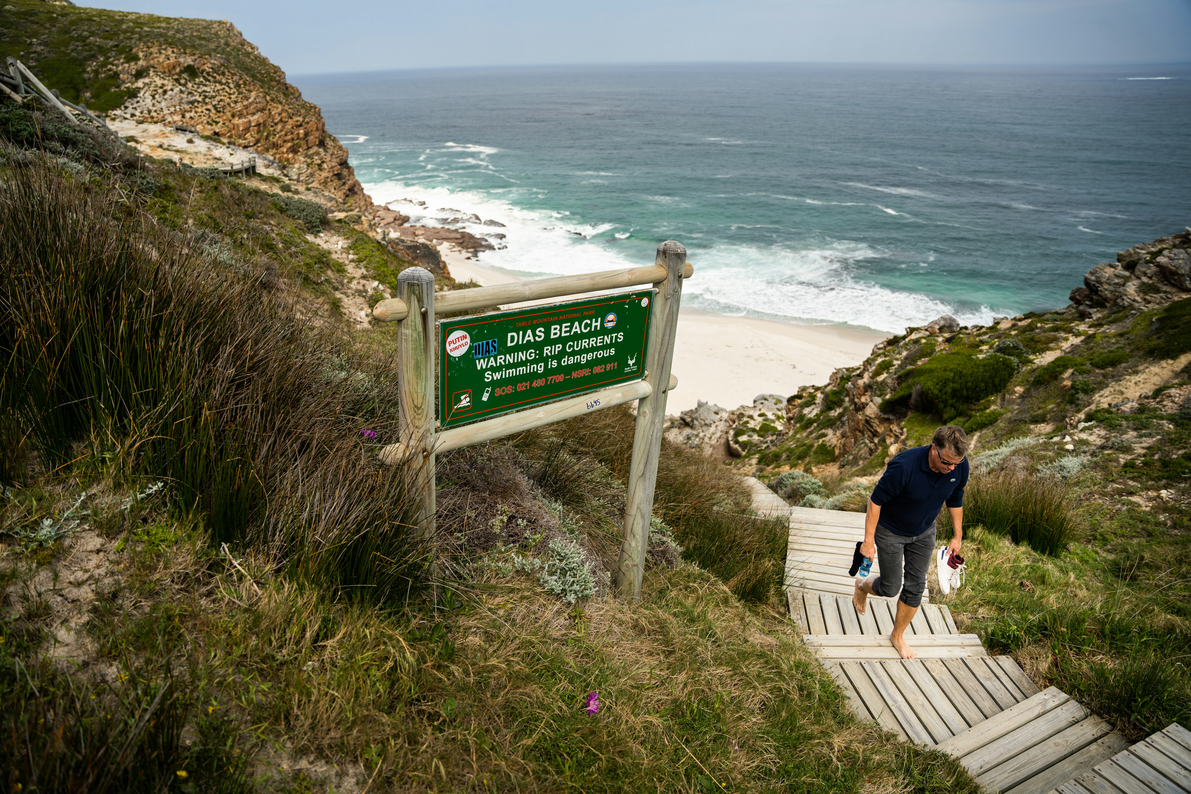 A barefoot man walks up wooden steps from a beach cove. Cliifs are visible on either side of the beach.