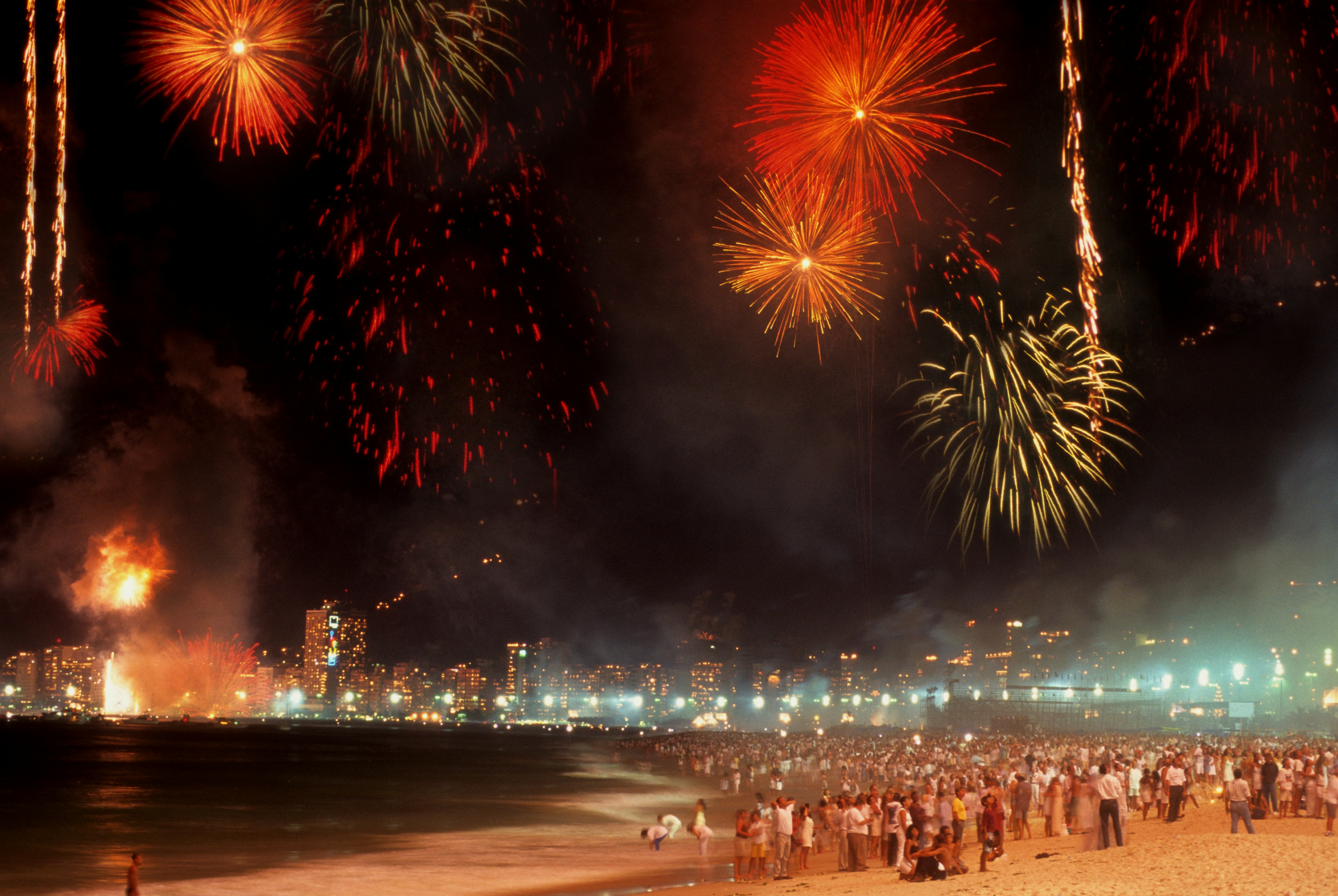 Colorful fireworks explode over a beach at night as thousands of people look on. The lights of buildings are visible on the shoreline in the distance.