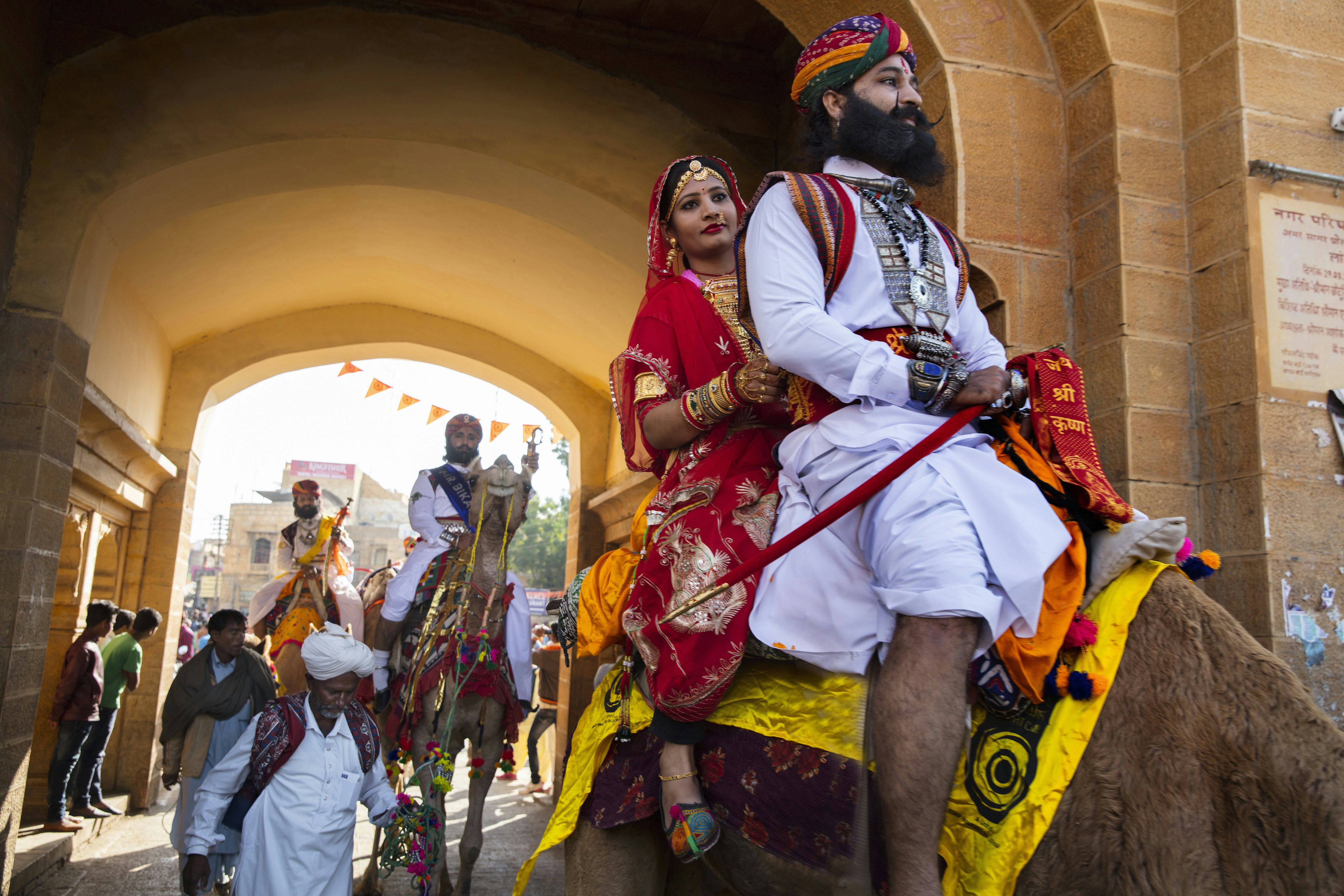 A man with a manicured beard and turban and a woman in a formal red sari ride a camel through an archway, followed by other people walking and on camels