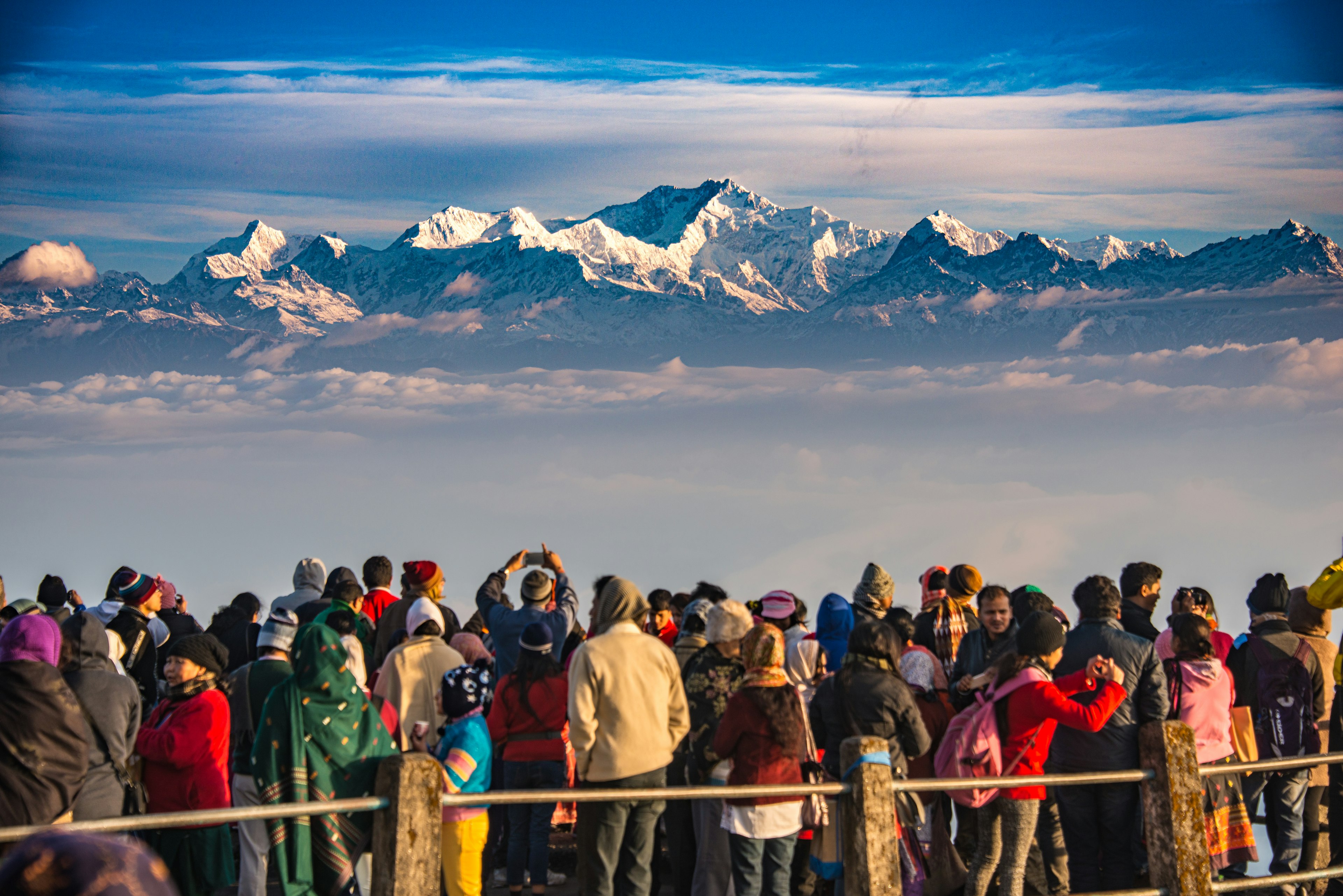 A large group of people gather at a viewpoint to watch the sunrise over a snow-capped mountain