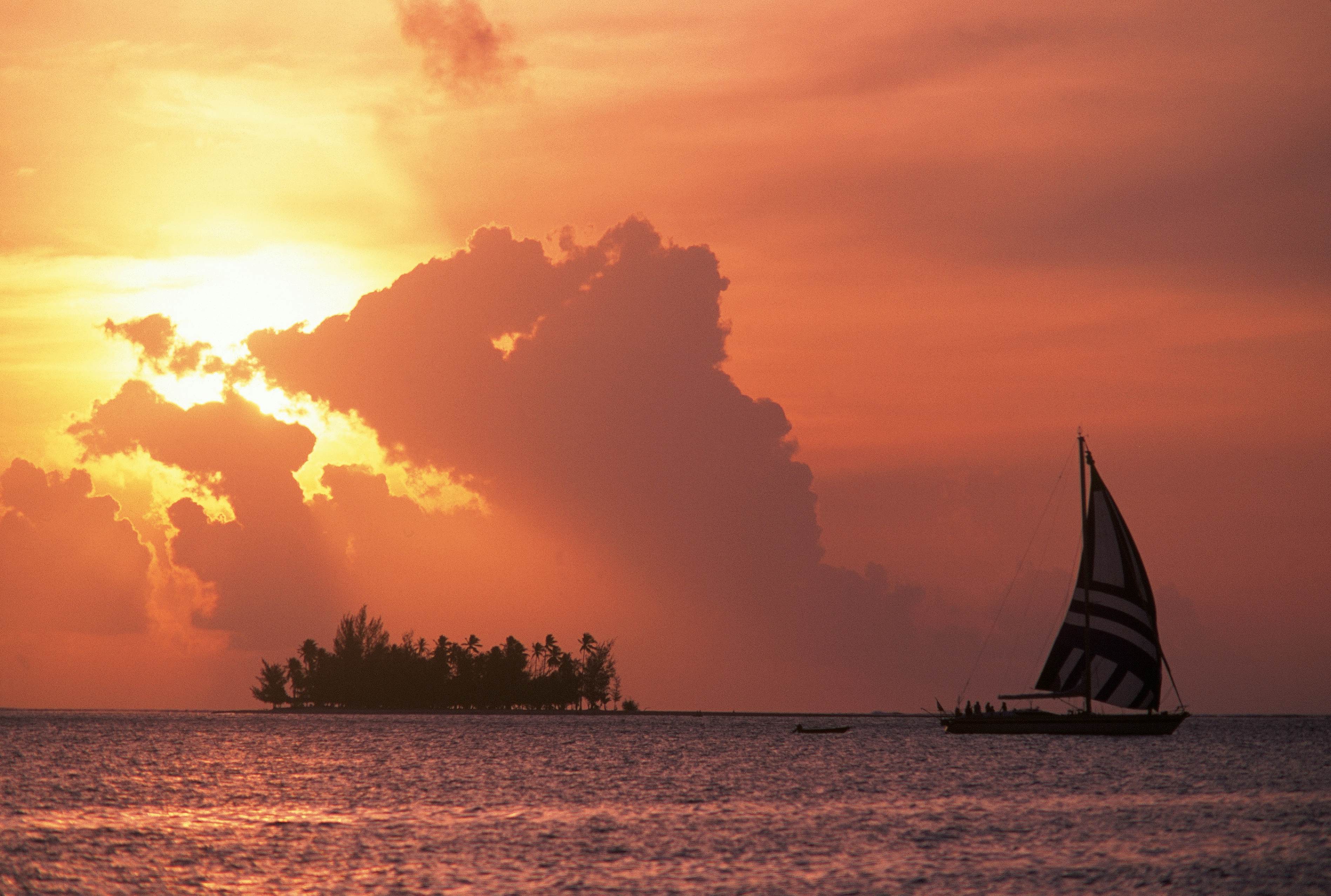 A sail boat out in a lagoon bathed in orange light as the sun sinks lower in the sky behind a large cloud