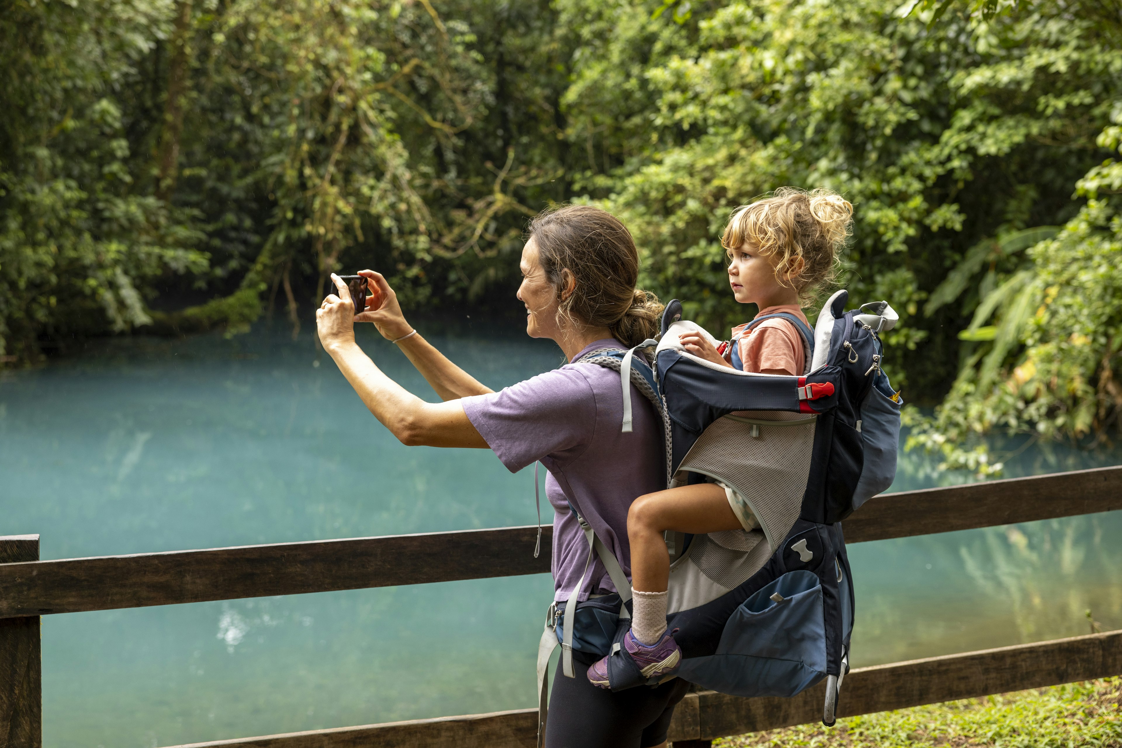 A woman taking a photo of a blue lagoon while hiking with her daughter, who is in a carrier on her back. Dense tropical vegetation surrounds the lagoon.