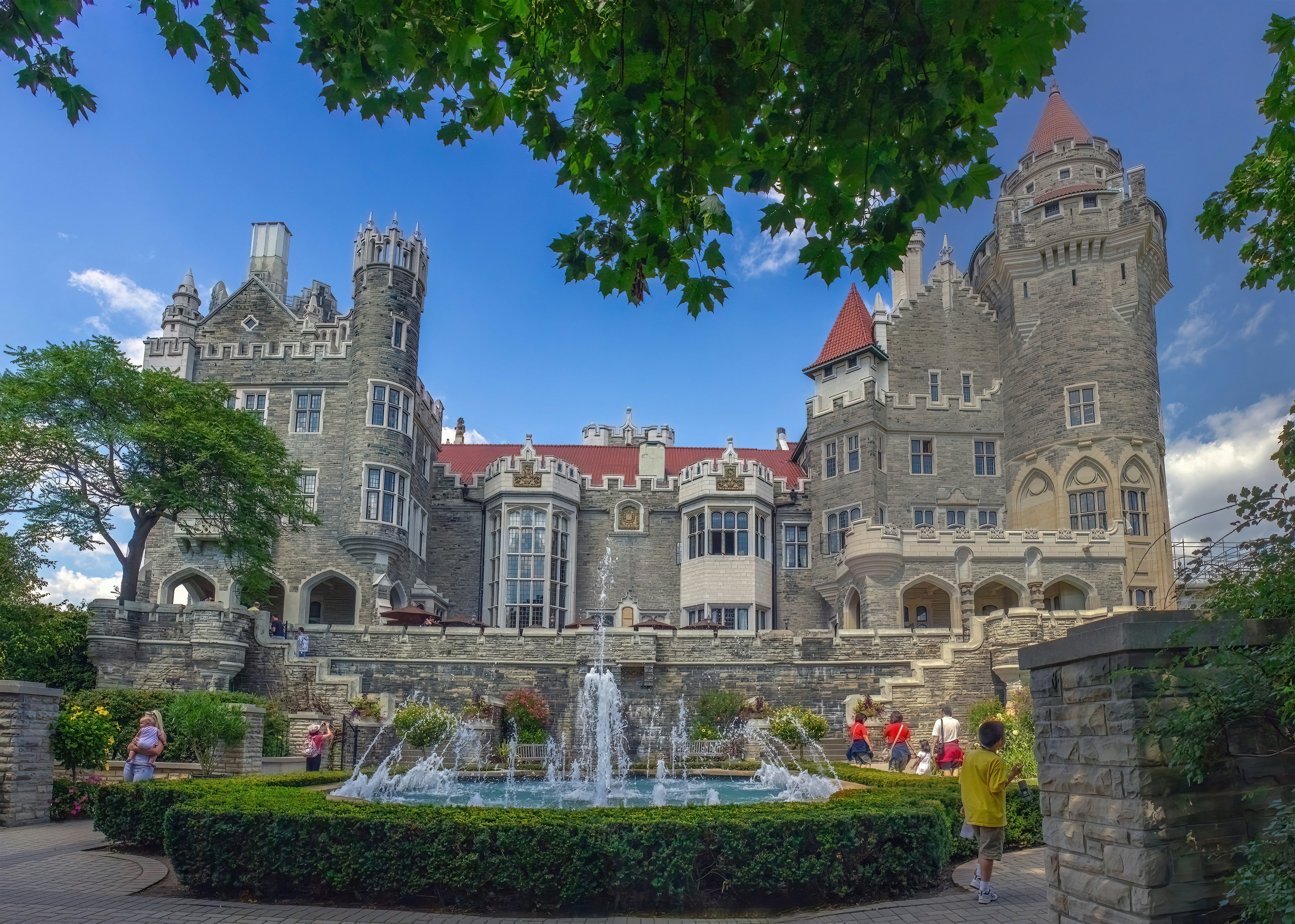 People walking by a fountain in the gardens in front of a large Gothic-style castle