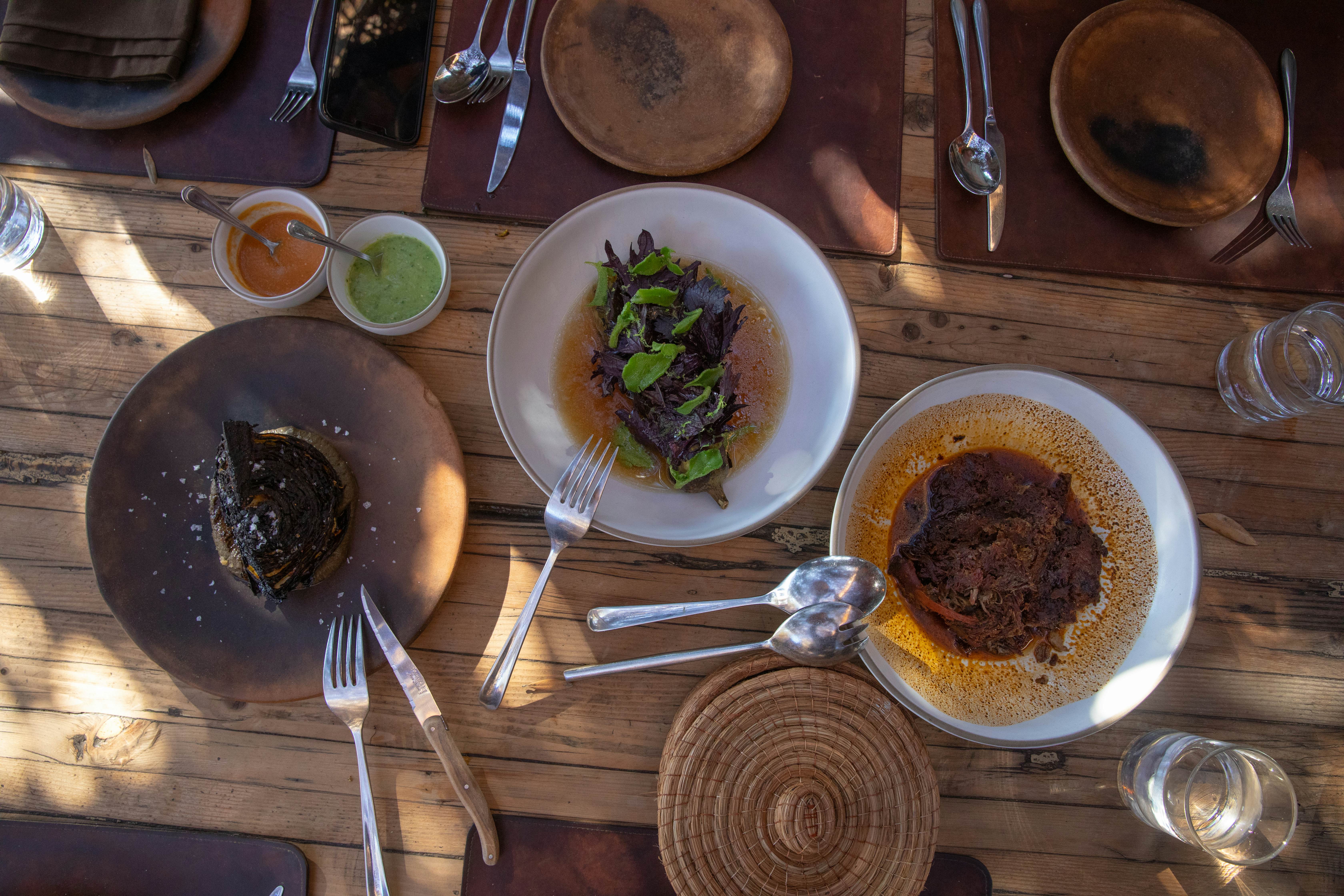 An overhead view of gourmet dishes arrayed on a wooden table with rays of sun coming through a pergola above