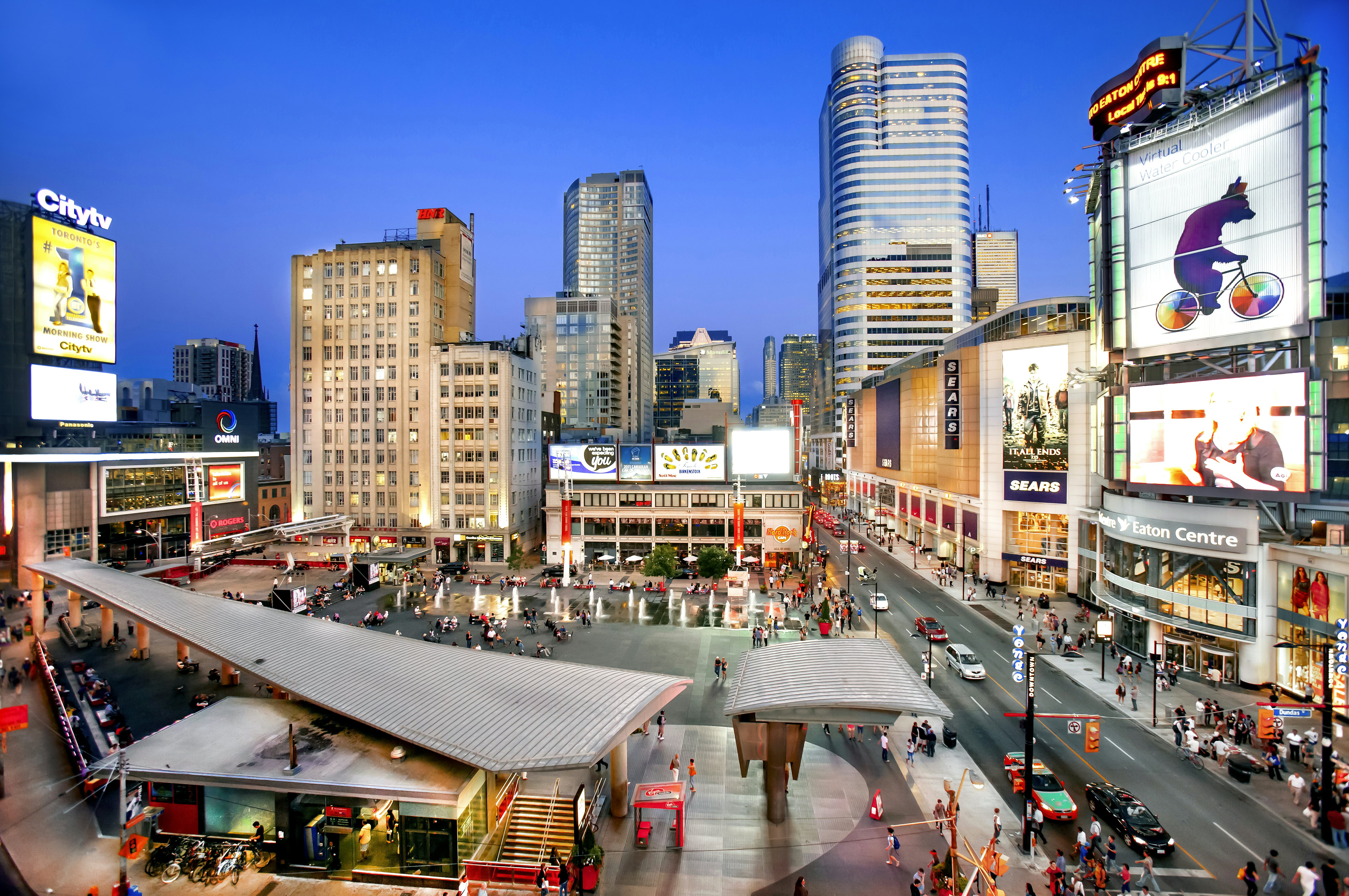 A brightly lit city square with TV screens showing advertising and people sat near pavement fountains