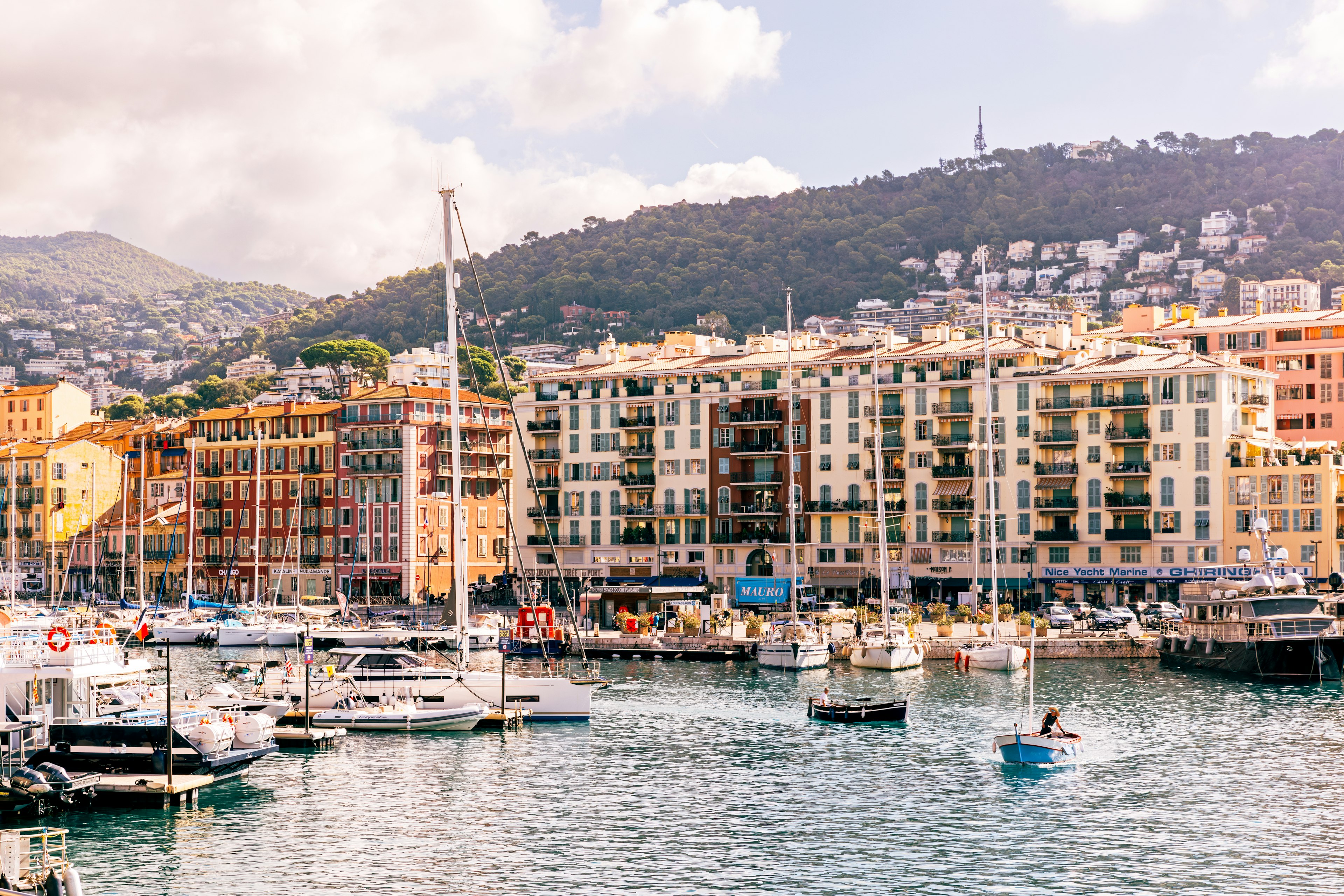 Sailboats are moored in a city port, with apartment towers along the waterfront and houses visible in the green hills beyond