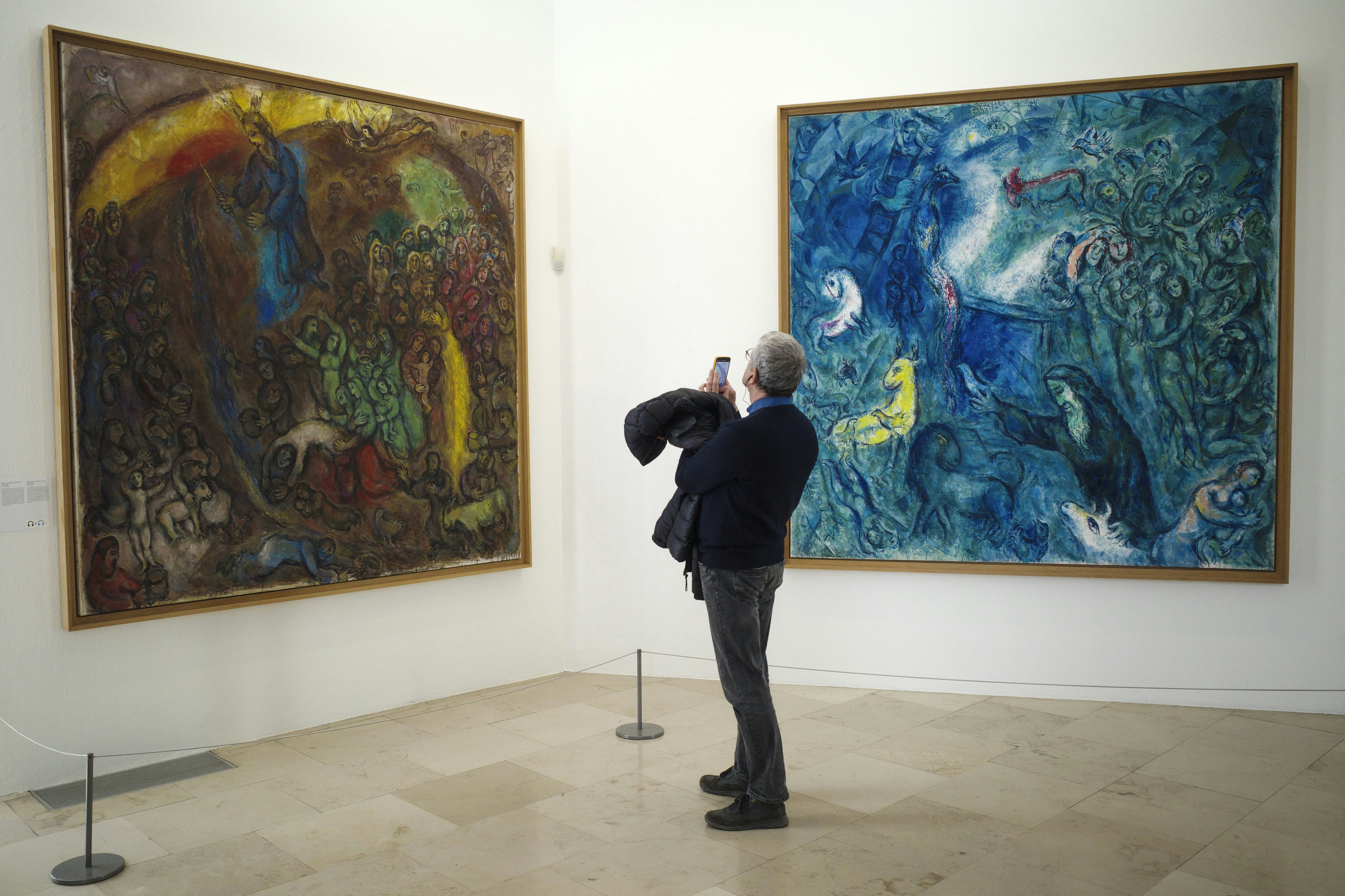 A man stands in a gallery and uses his phone to take a photo of an artwork. Two large paintings – one in red, gold and other colors; the other in mostly blue tones – hang on the white walls.