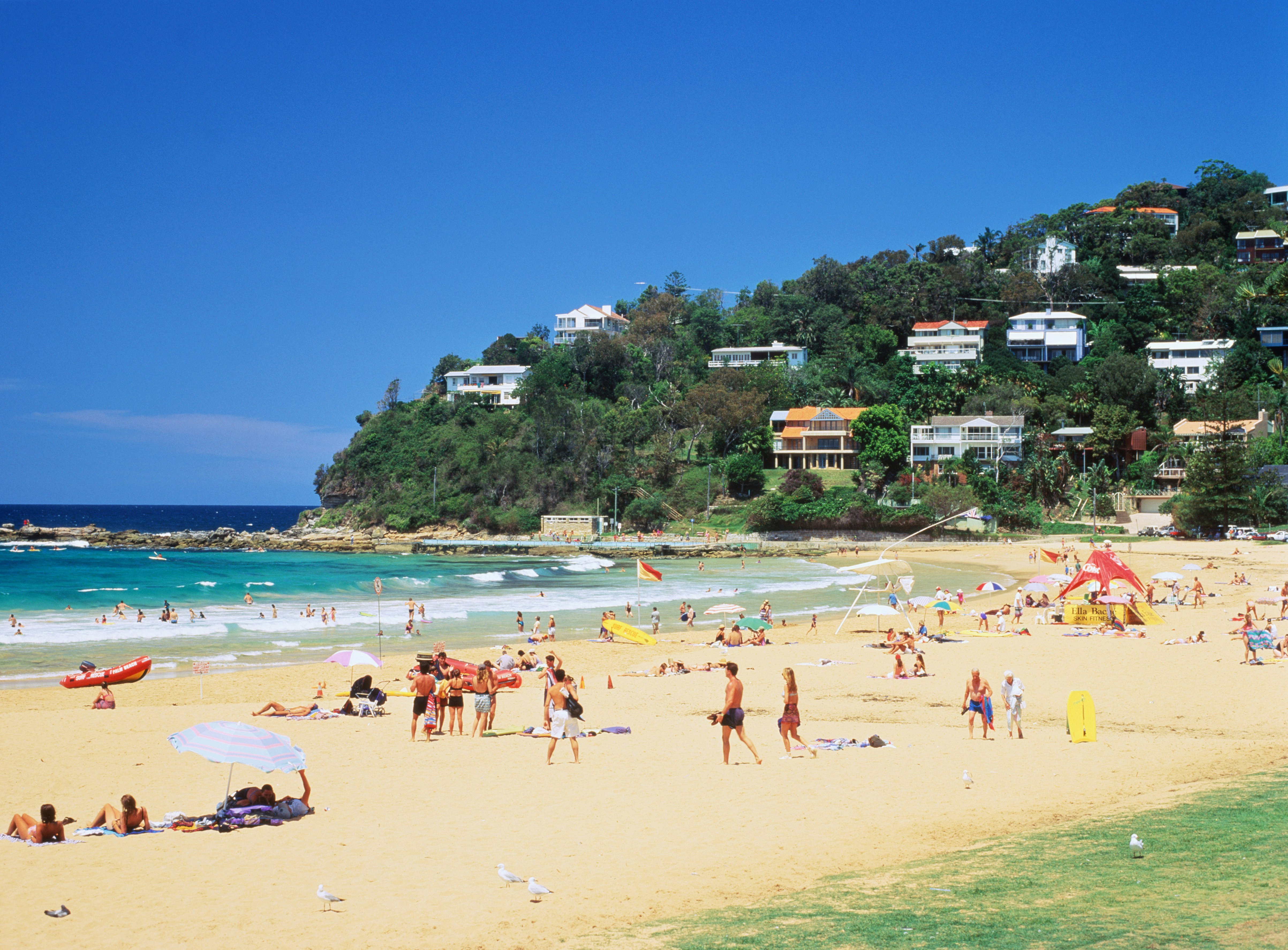 People on a beach – some lying out in the sun, some playing games, some swimming in the surf – in a cove with gentle waves, and a green slope with houses