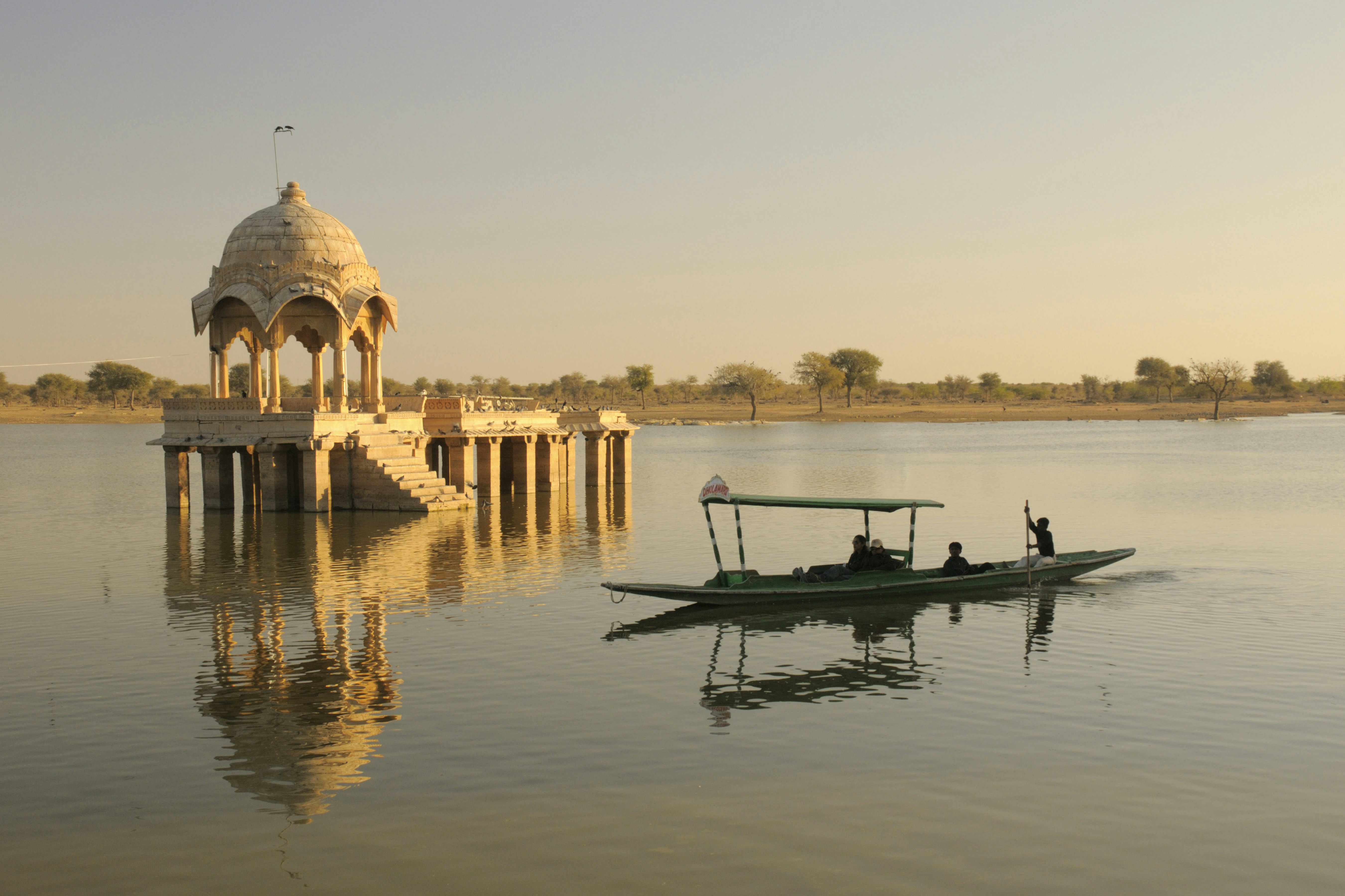 A person uses a pole to propel a long boat with a canopy past a monument on an island with columns and a dome on a lake at sunset