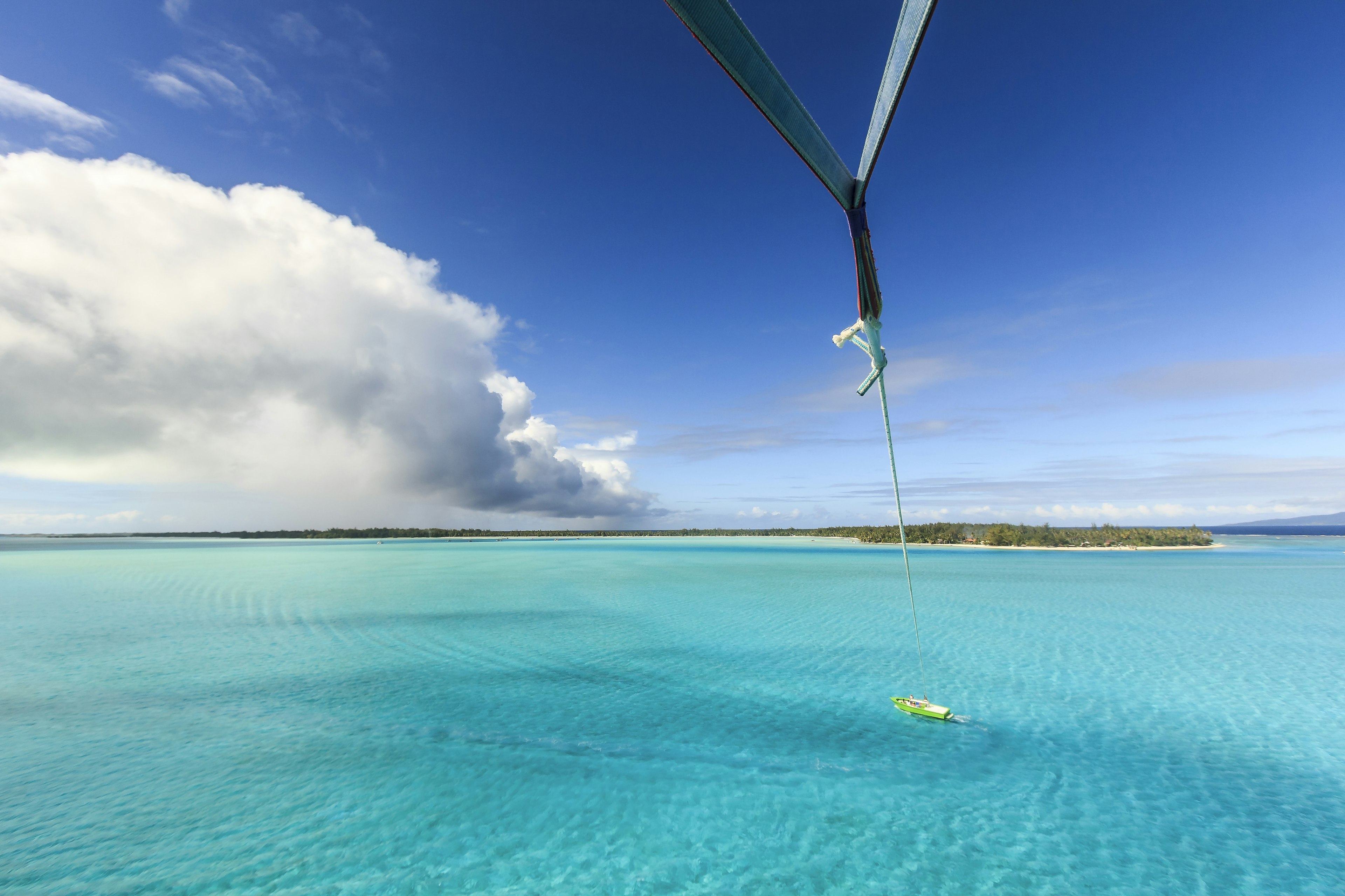 An overhead shot taken by a parasailer in the air. Blue waters and the boat attached by cord to the parasail, as well as green islands, are visible.