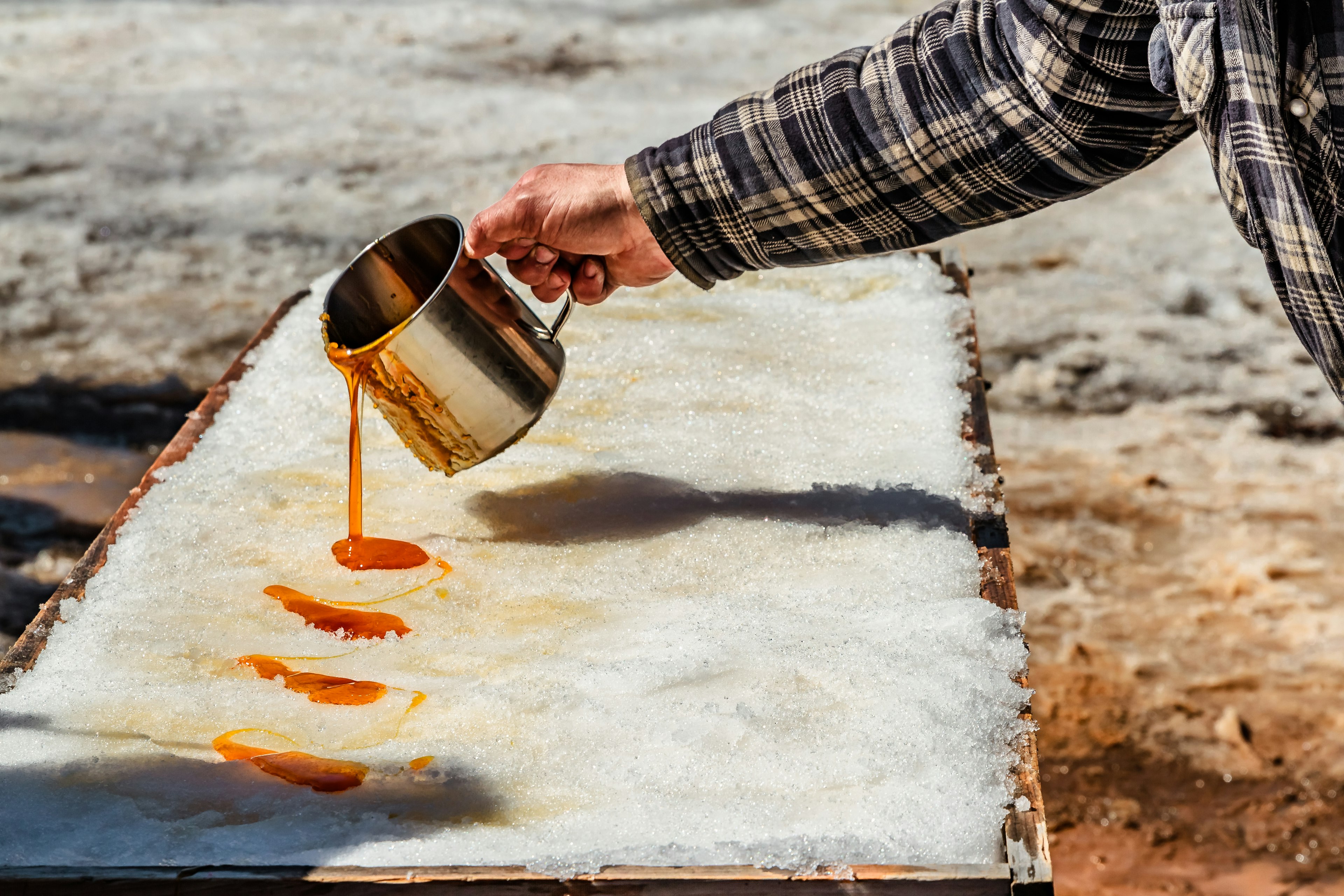 A person in a flannel shirt pours maple syrup onto snow.