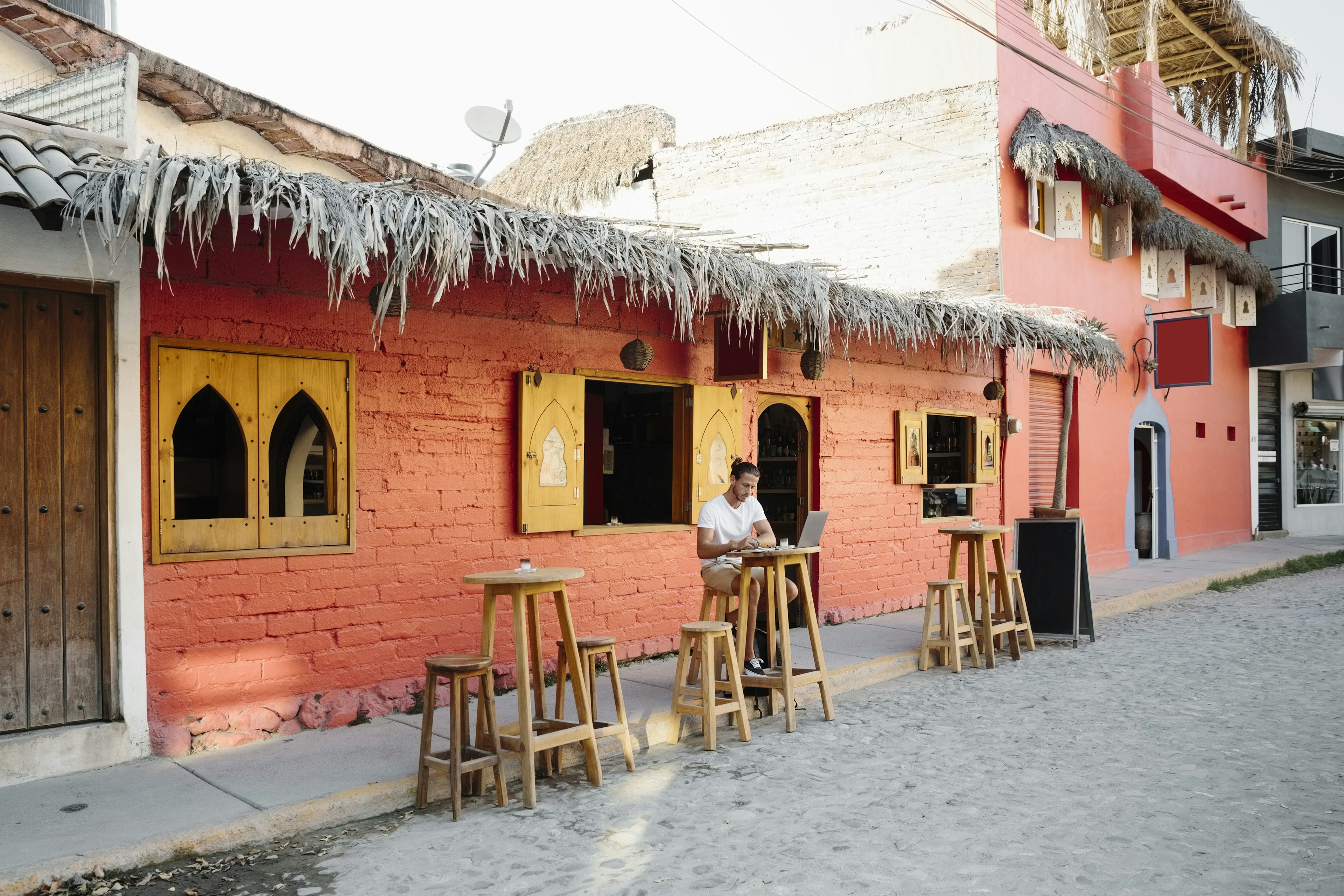 A solo traveler sits at a bar stool outside a cafe typing on a laptop