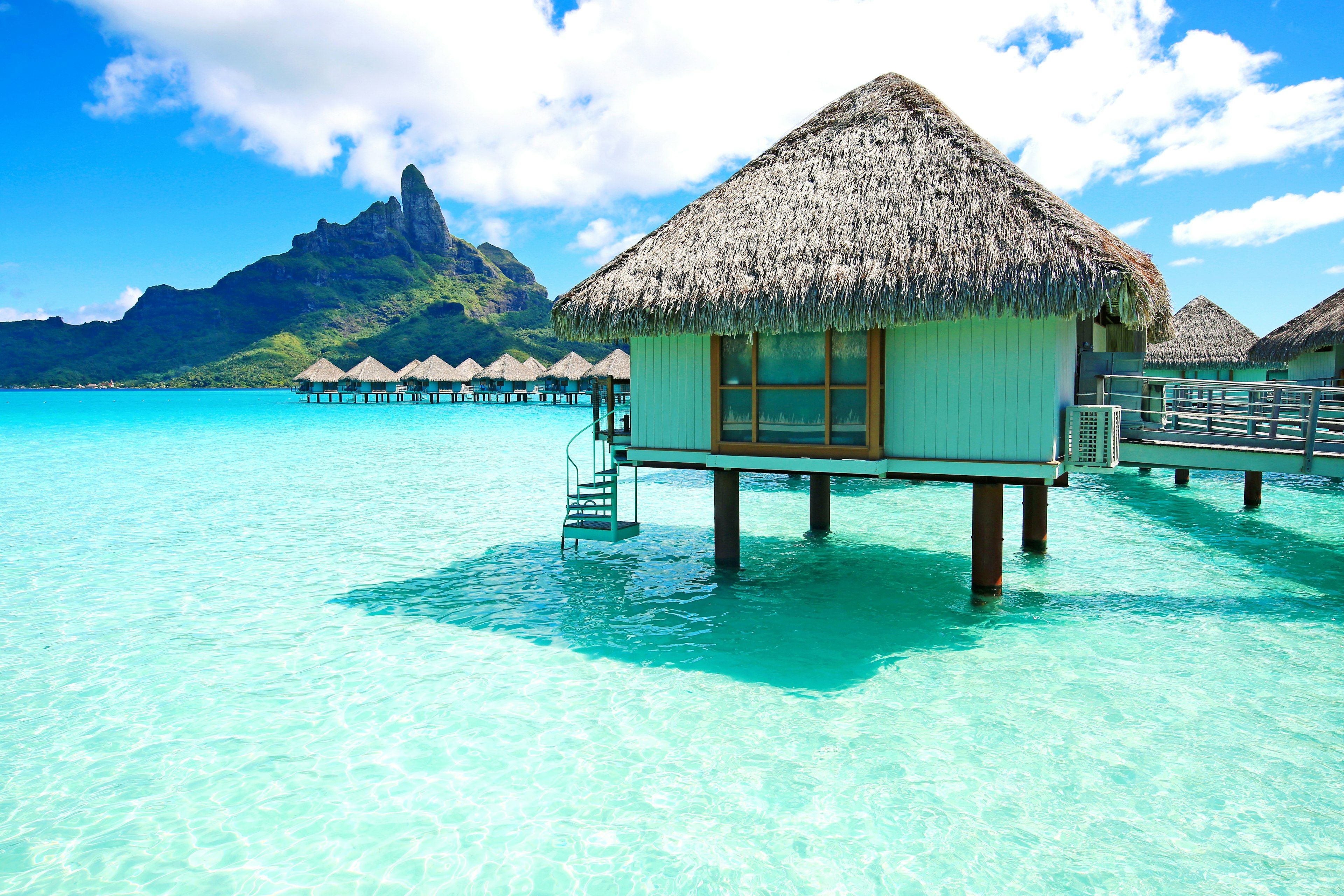 A bungalow with a thatched roof sits on pilings set into the azure waters off a tropical island. A green mountain is visible on shore.