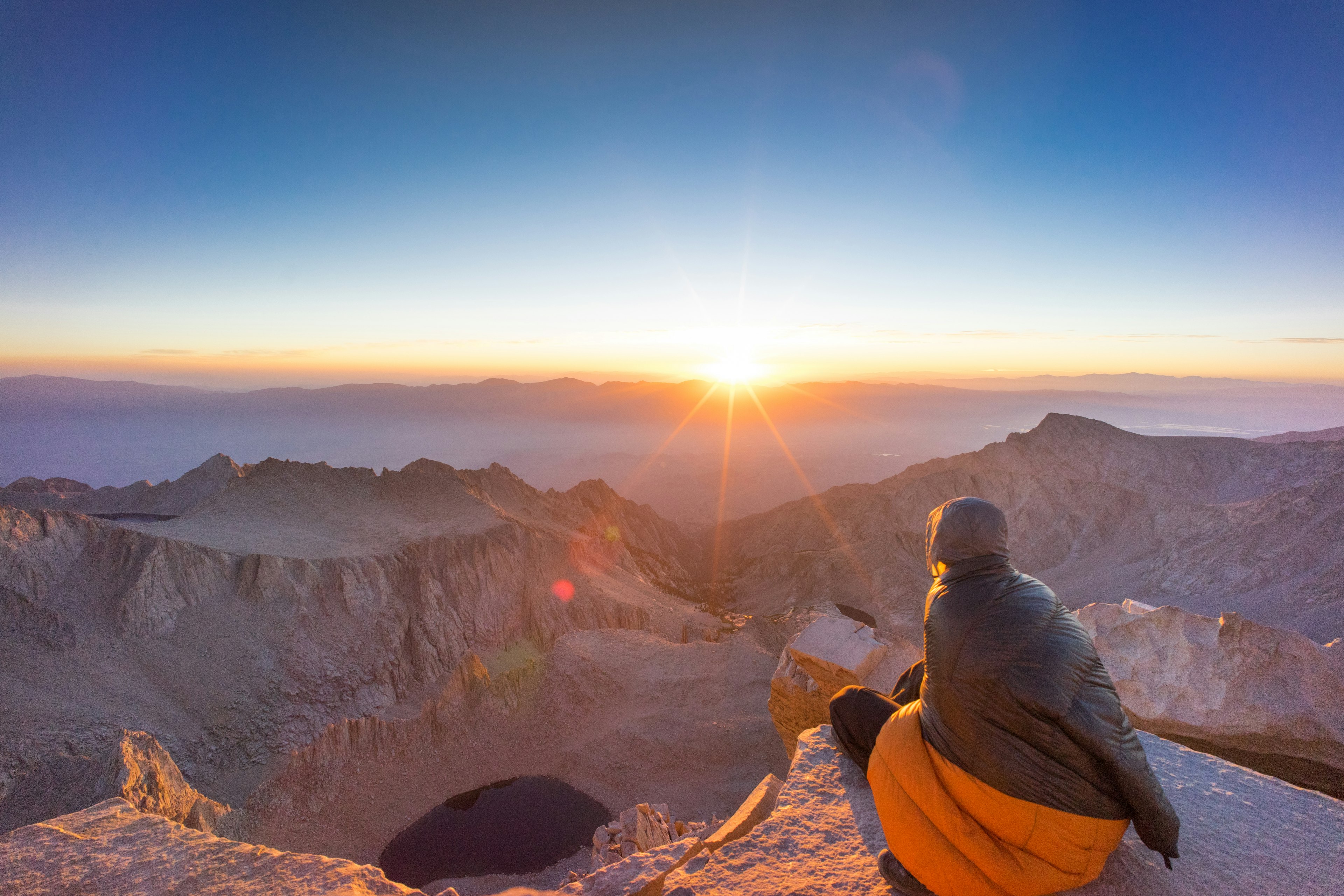 A crouched hiker watches the sunrise on the top of Mount Whitney.