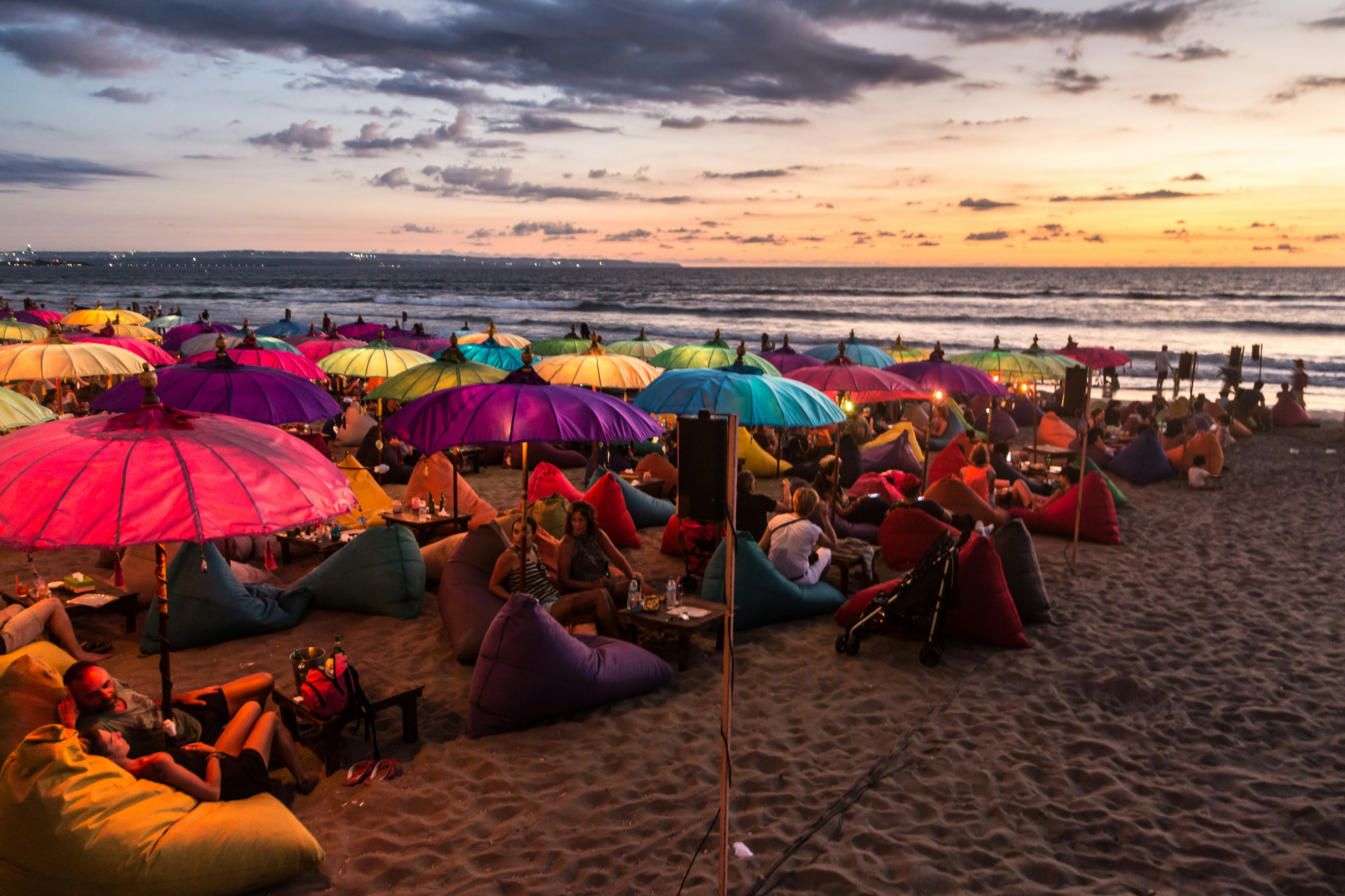 A crowd of people share food and drink under colourful umbrellas during sunset on a beach