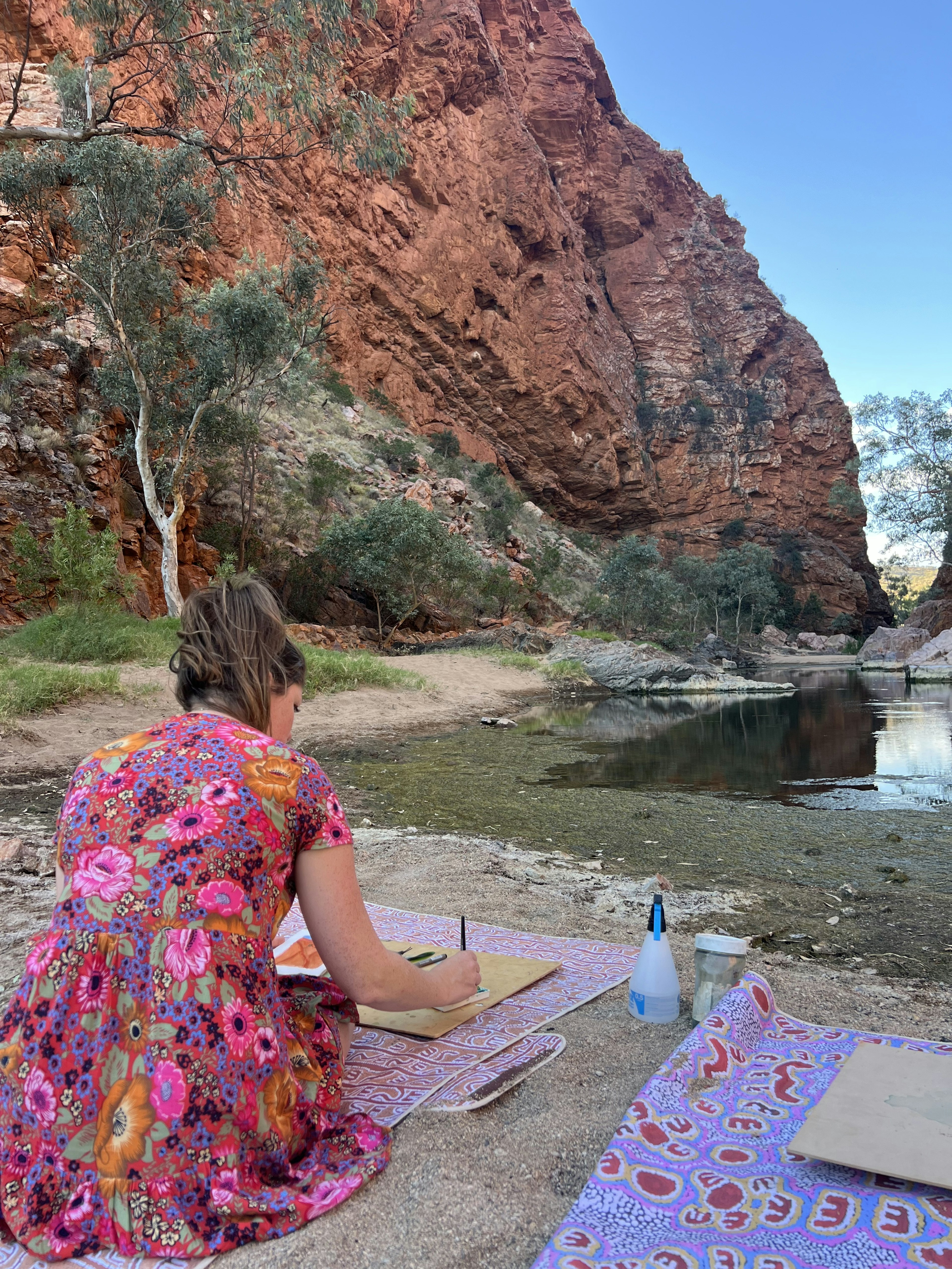 The writer kneels on the floor to work on a painting at a waterhole in the Outback