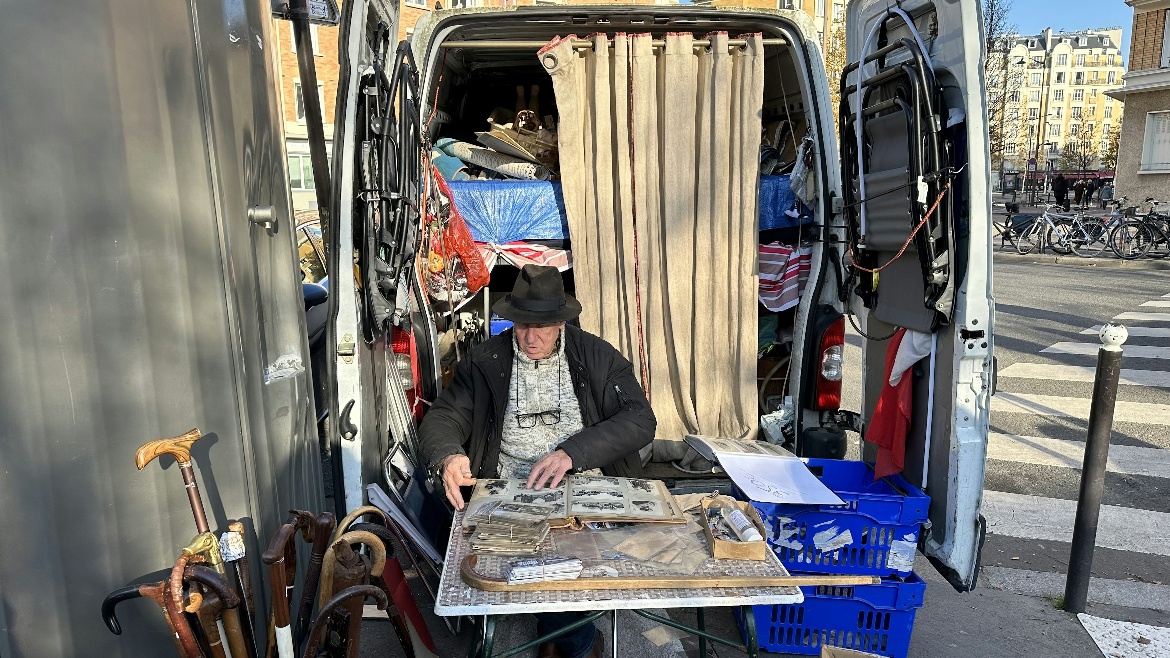 A market trader sits at a table at the back of a van that's packed with items for sale