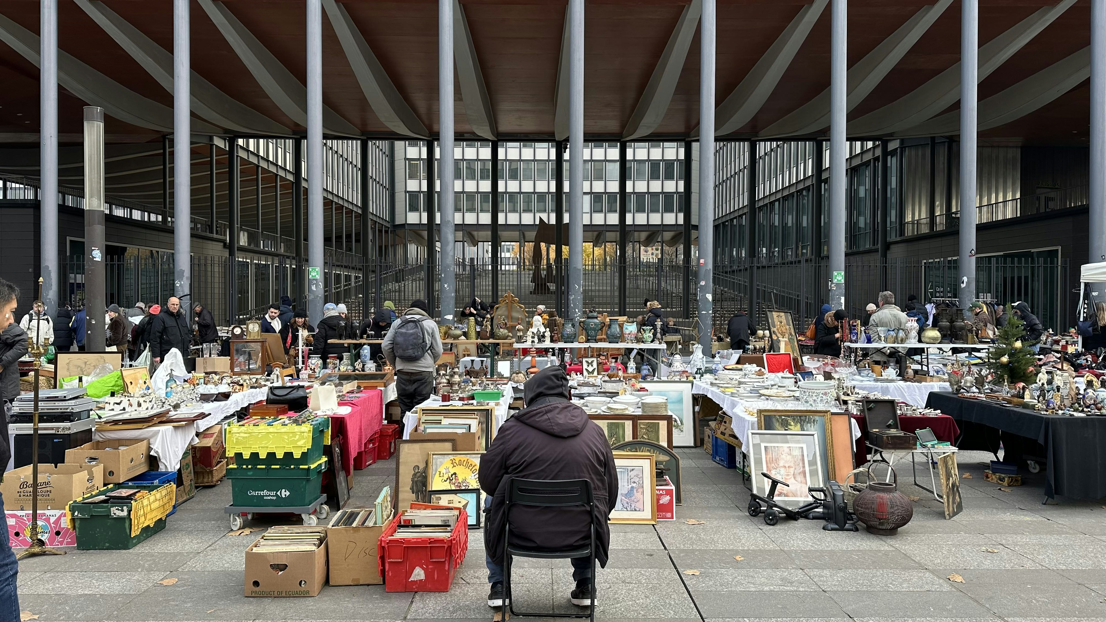 A vendor sits on a low stall in front of a series of market stalls selling paintings, vases, ornaments and other decorative items