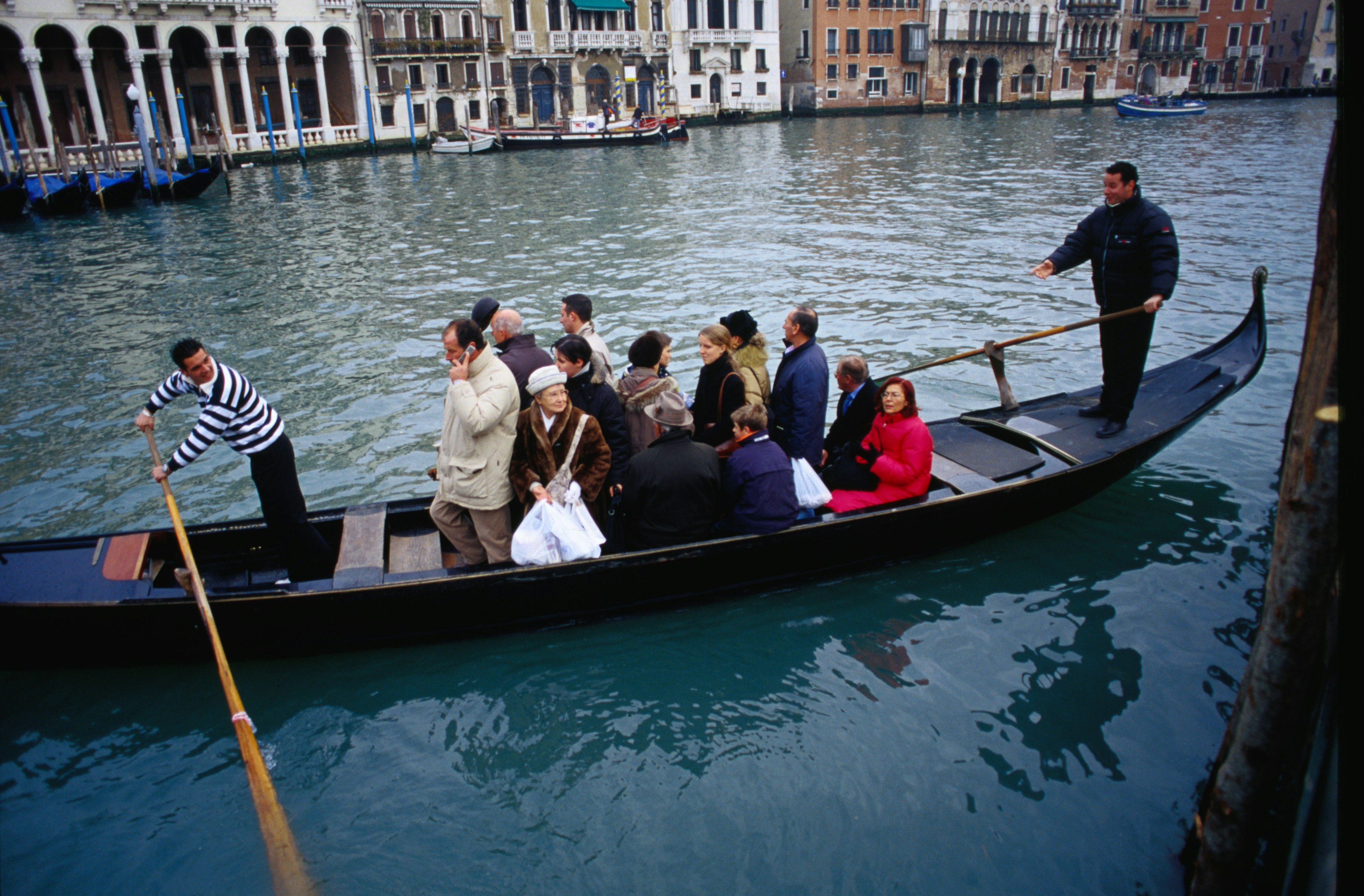 Gondoliers push off a dock, their boat filled with passengers crossing a canal