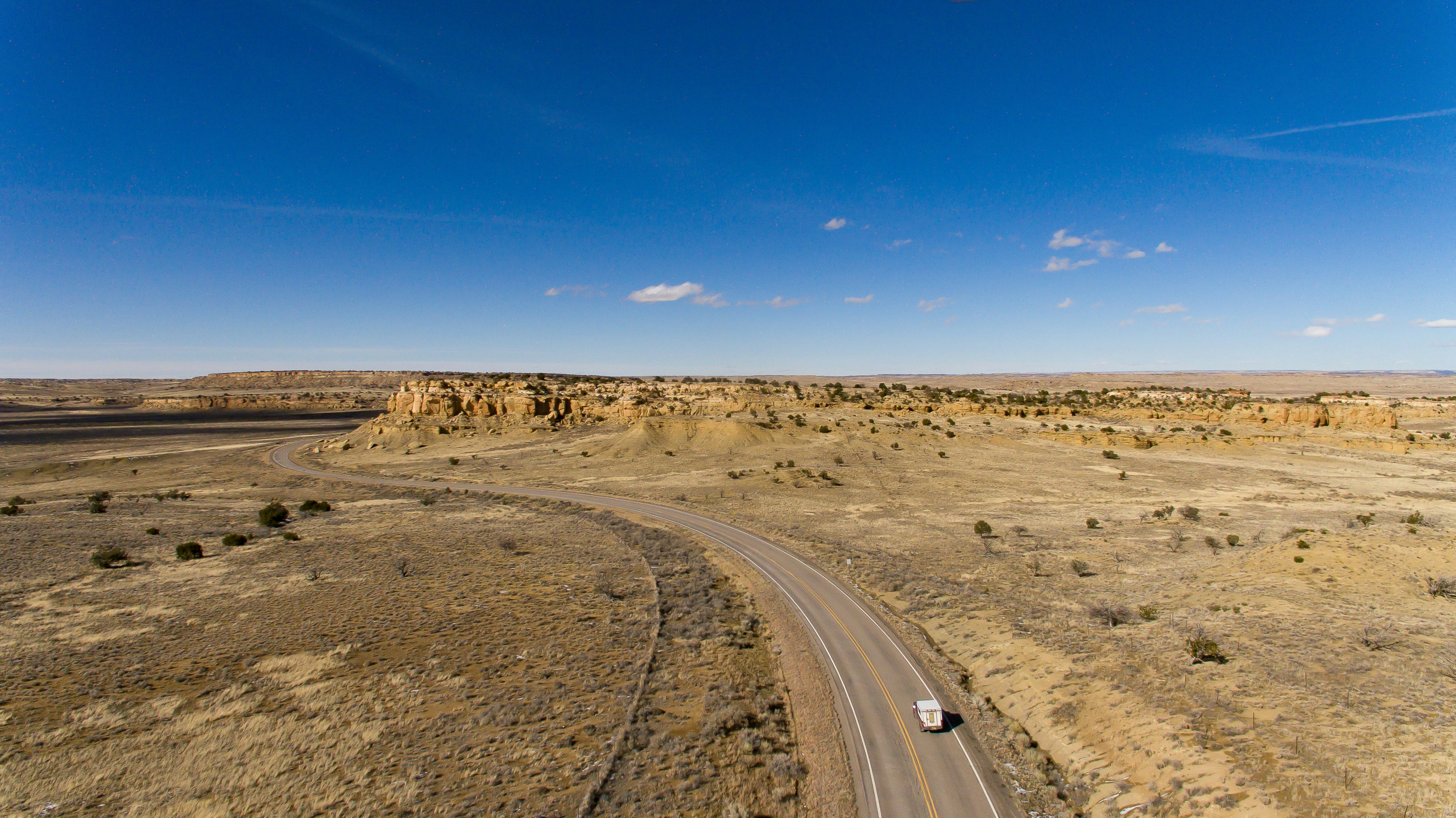 An aerial view of a lone camper on a winding road through the desert. Flat-topped mesas and a blue sky are visible in the distance.