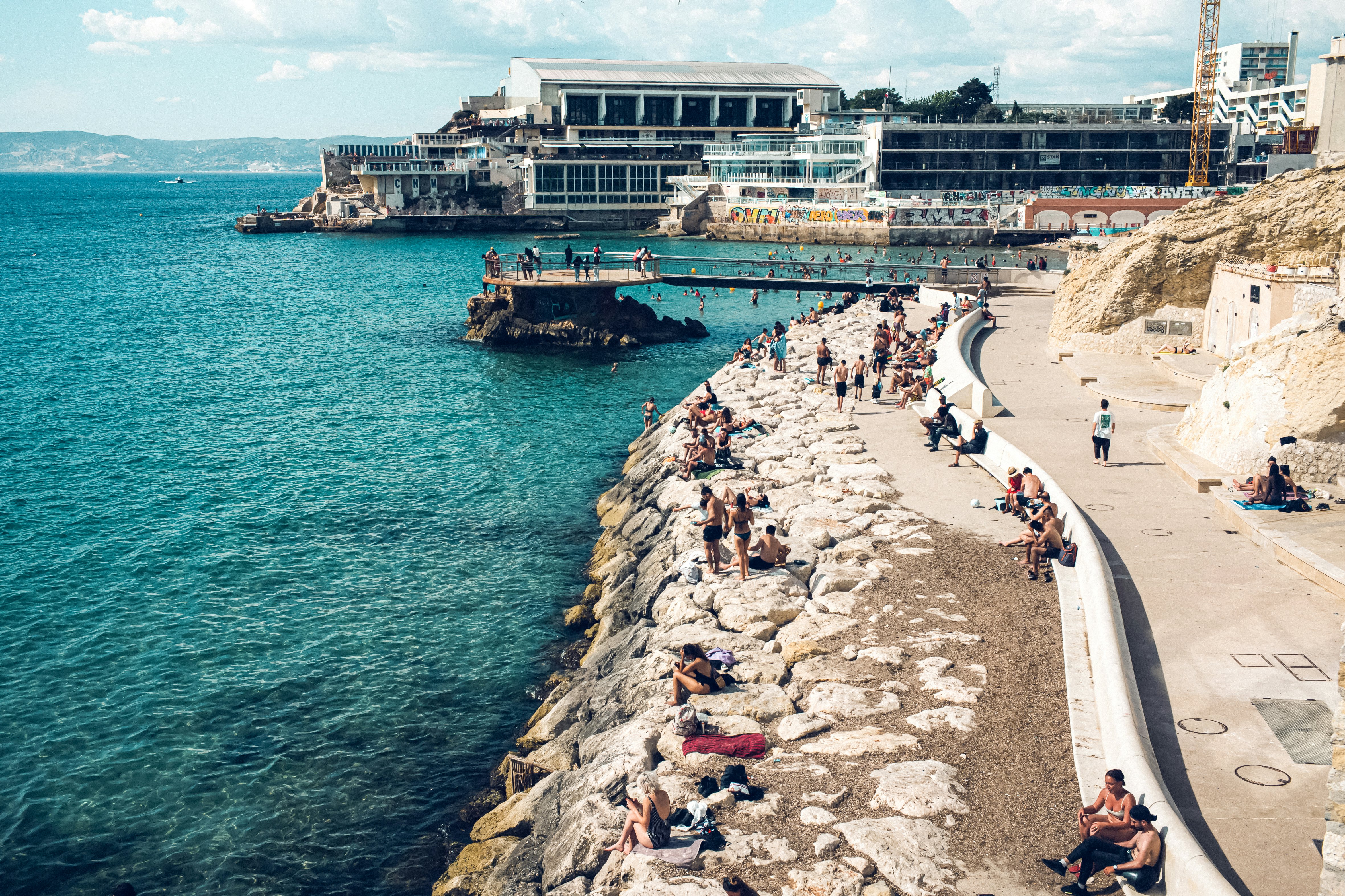 People lounge on rocks on a pathway beside the blue sea, with hotels and restaurants visible in the distance