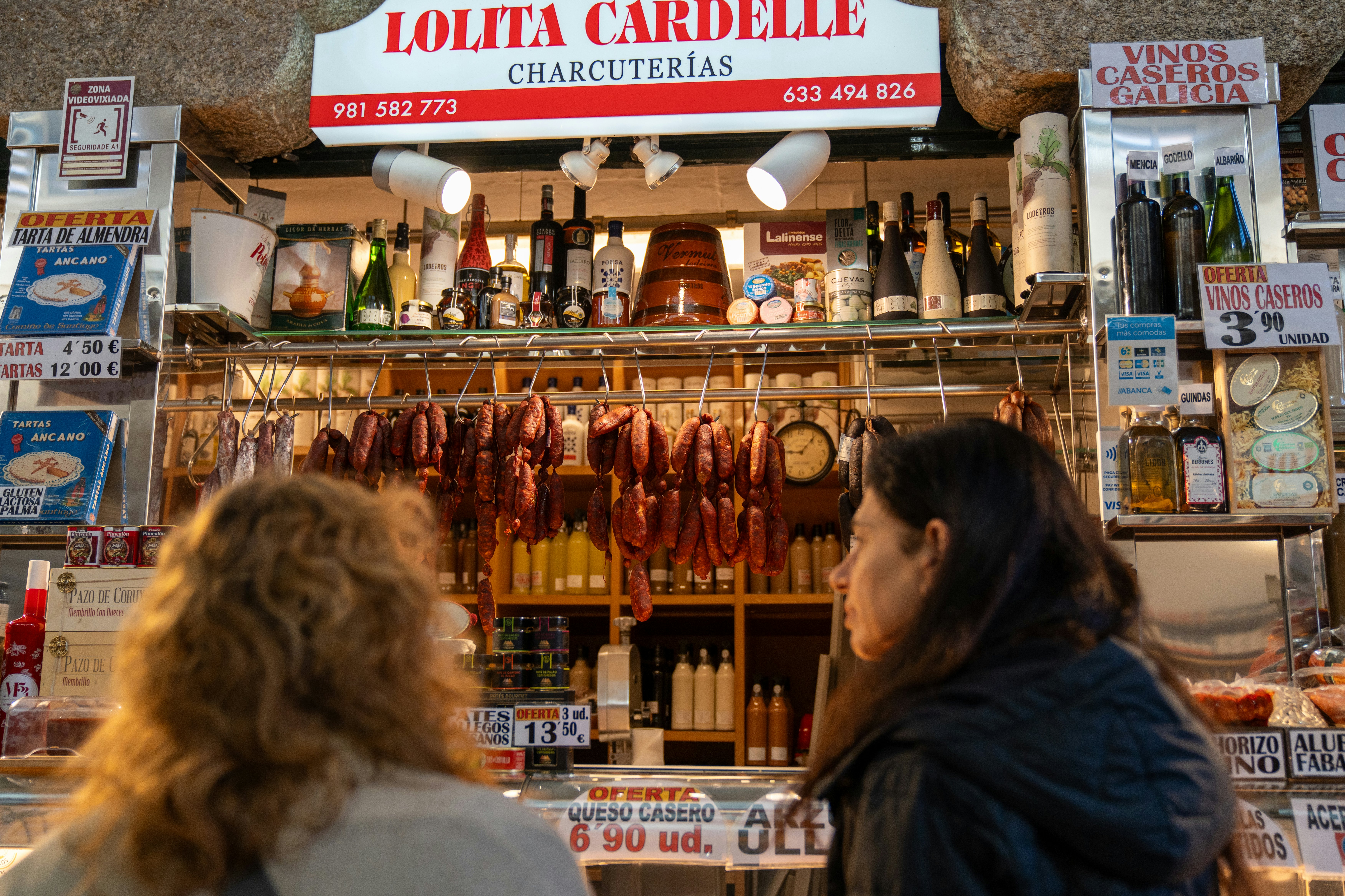 Two people at a counter with wares on shelves and sausages hanging behind the counter.