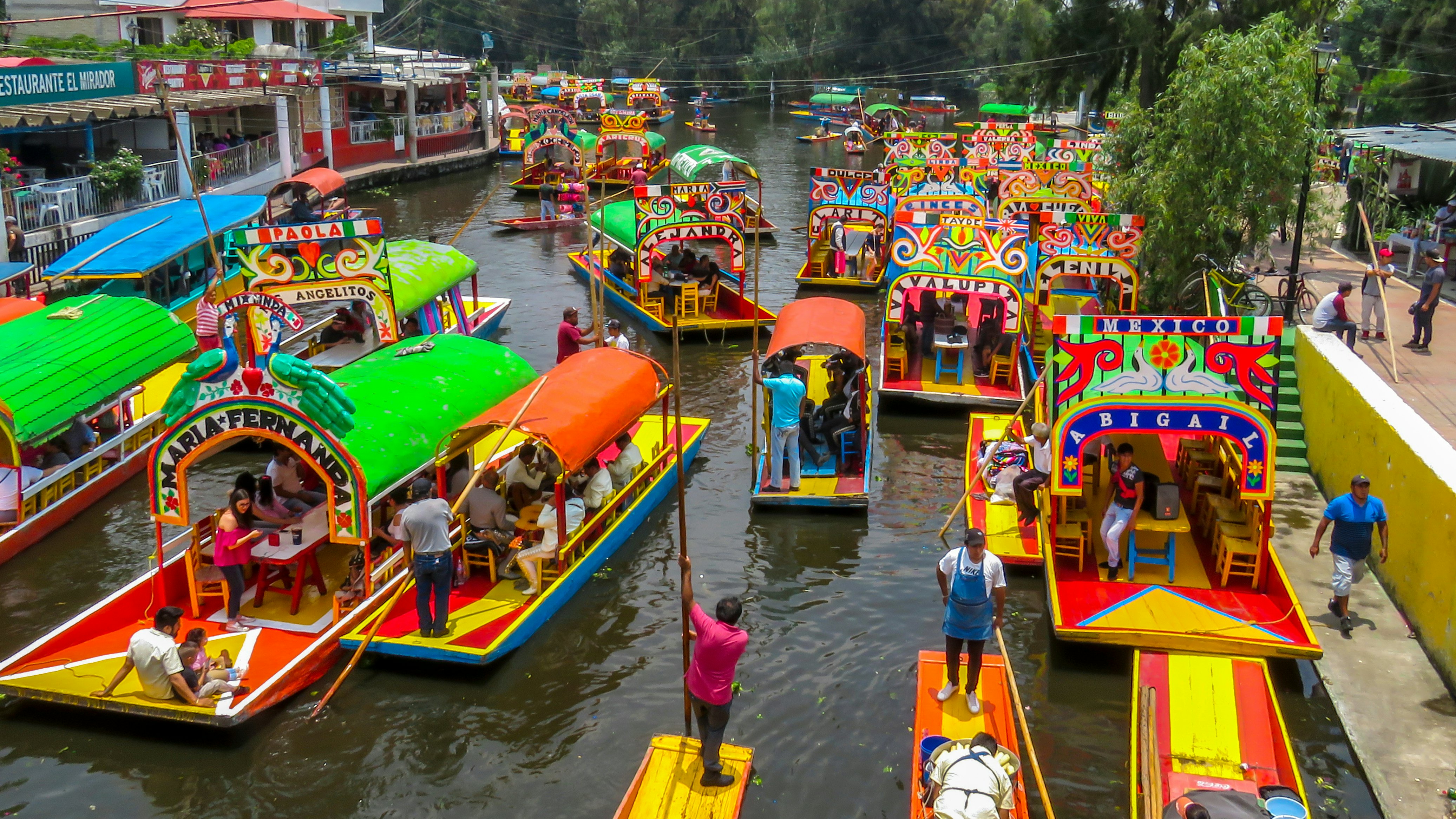 An overhead view of a flotilla of brightly colored barges on a canal in a city.