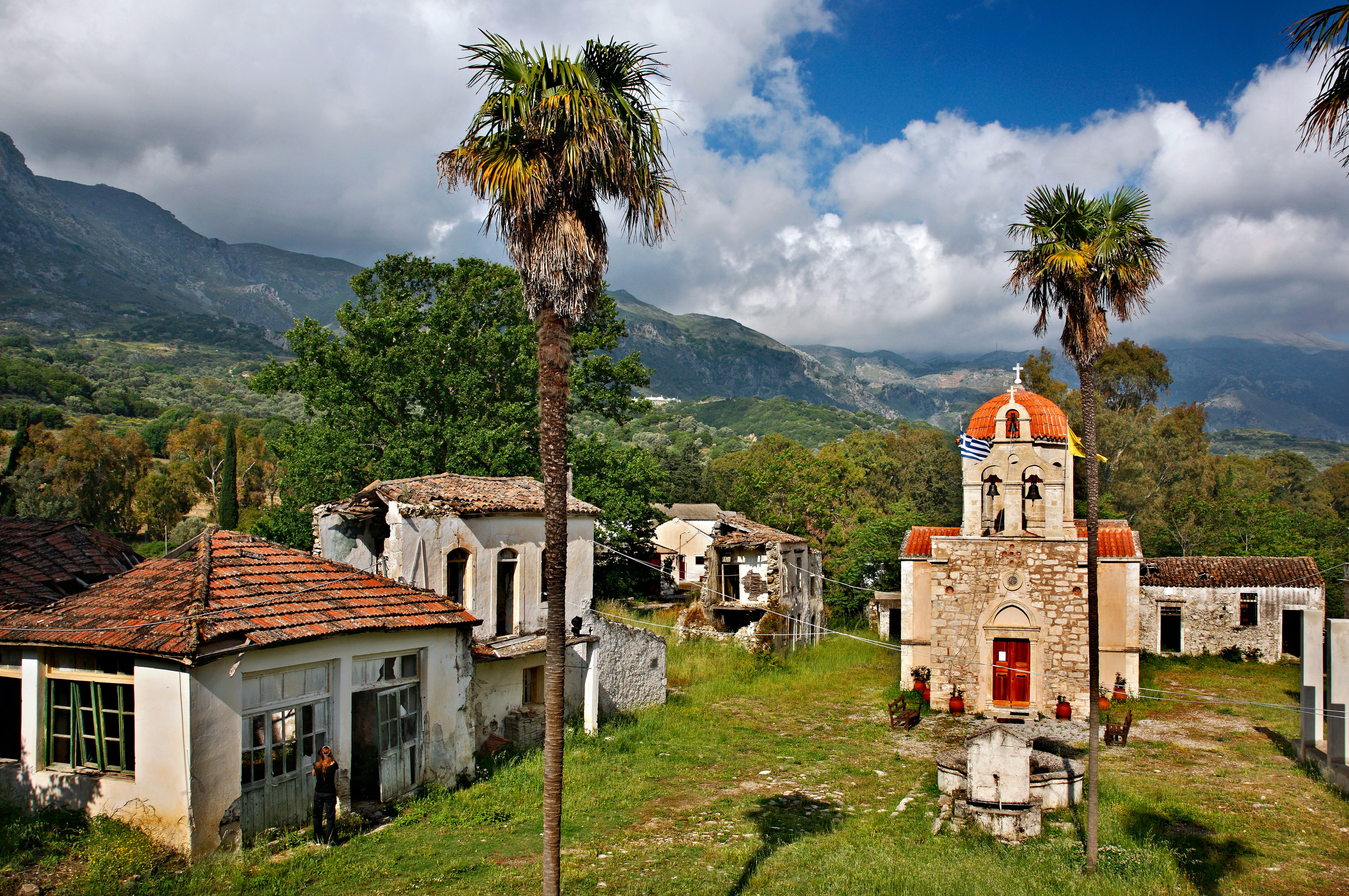 An abandoned Greek monastery sits in the hills with tropical trees in the foreground.