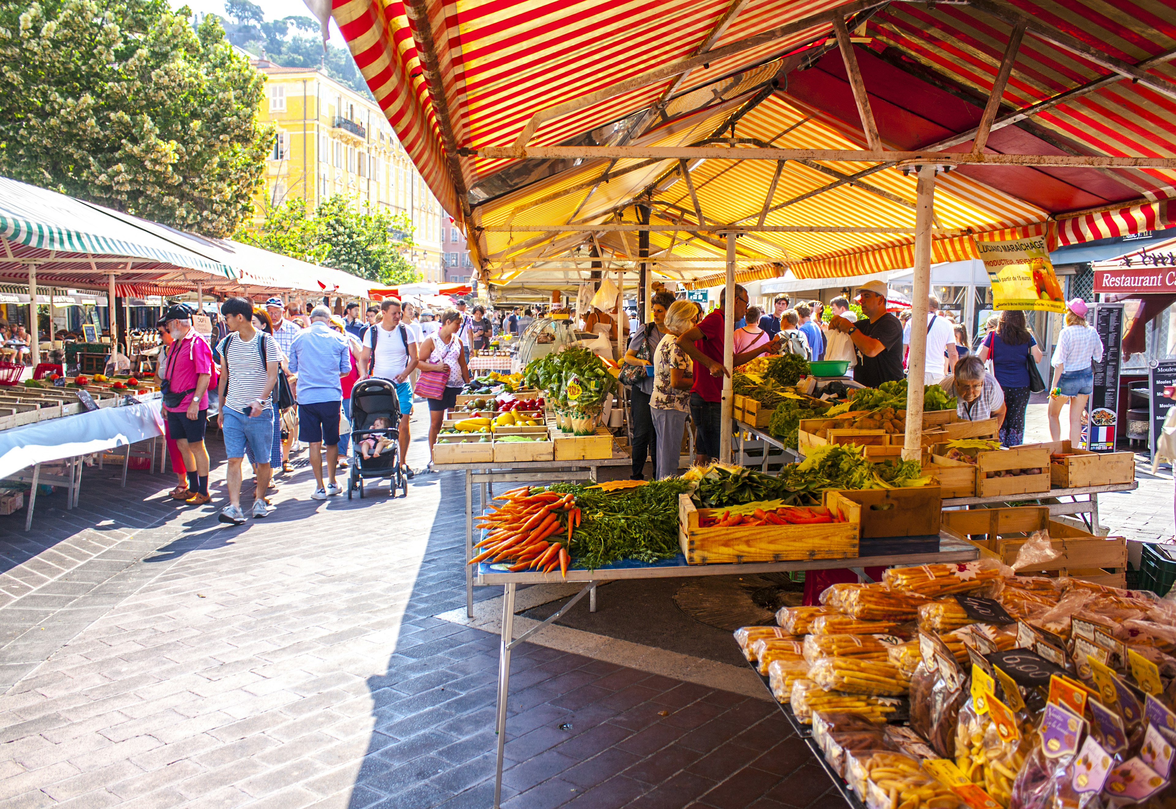 Various vegetables and fruit are laid out on counters of stalls at an outdoor market in the day, covered by awnings. People mill about in the background.