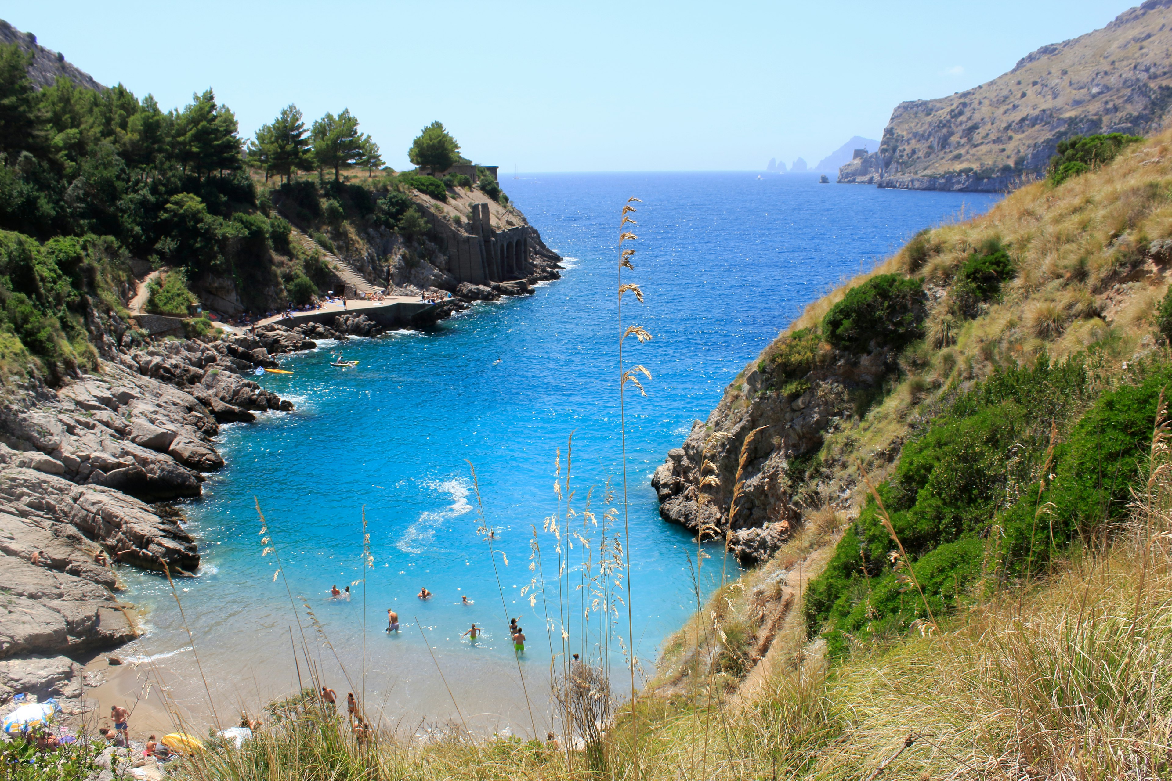 People swim in a deep blue bay, surrounded by trees and shrubs.