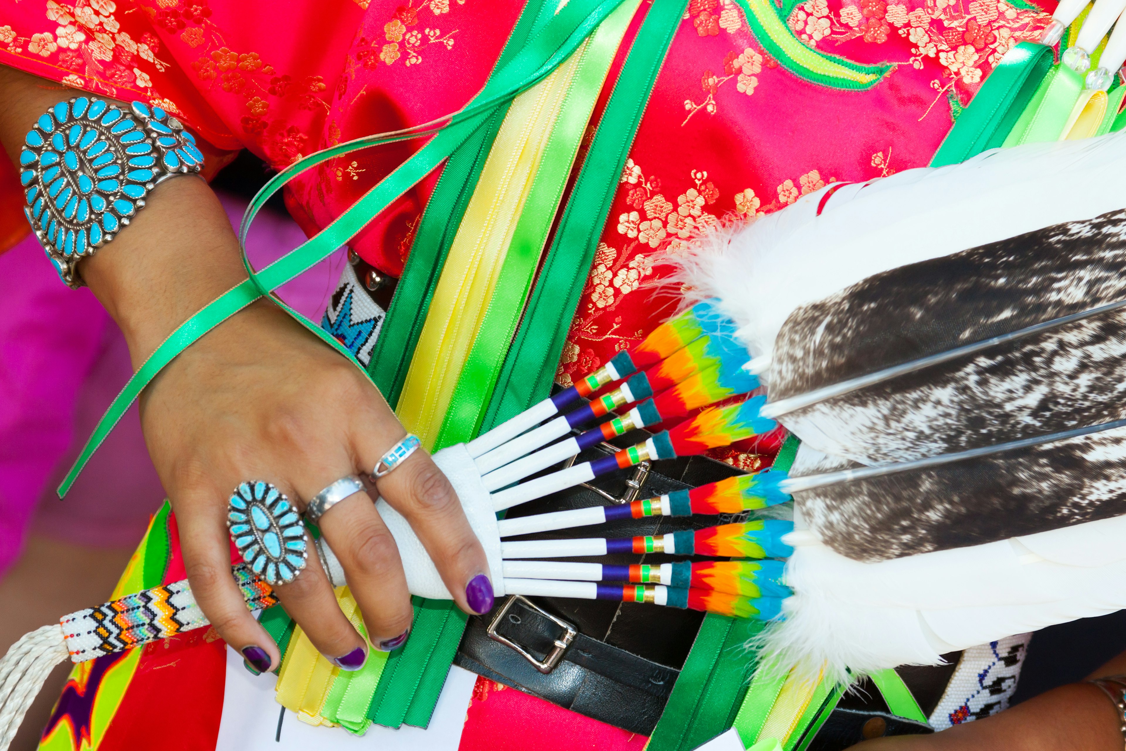 A close-up of a person holding a feather fan with bands of multiple colors while wearing an elaborate turquoise ring and bracelet and red and gold patterned clothing with green and yellow ribbons at the Santa Fe Indian Market fashion show