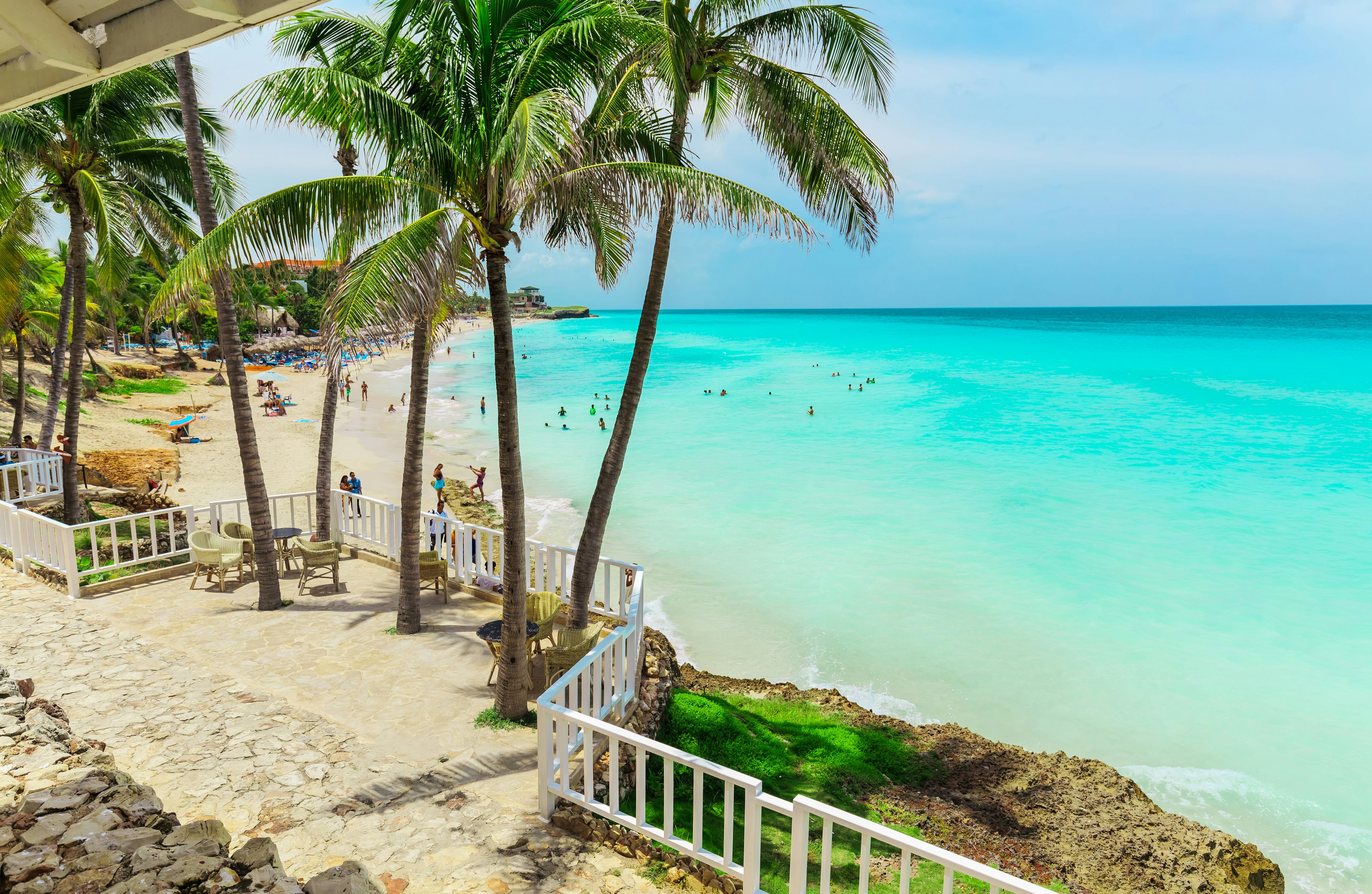 Swimmers play in the turquoise ocean while people relax on white sands of a palm-lined beach