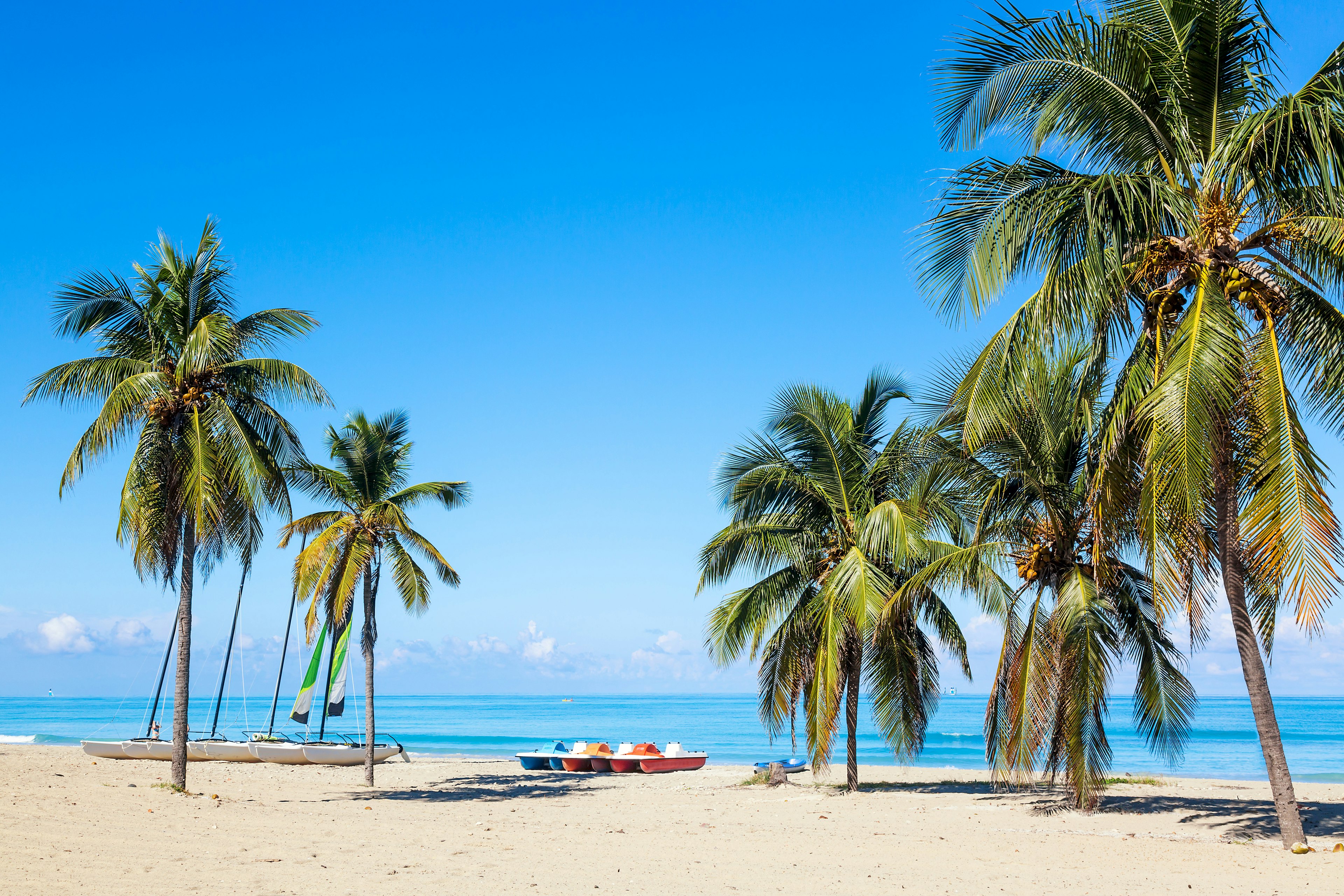 Several small boats sit on white sand between two clusters of palm trees on a beach in Varadero, Cuba