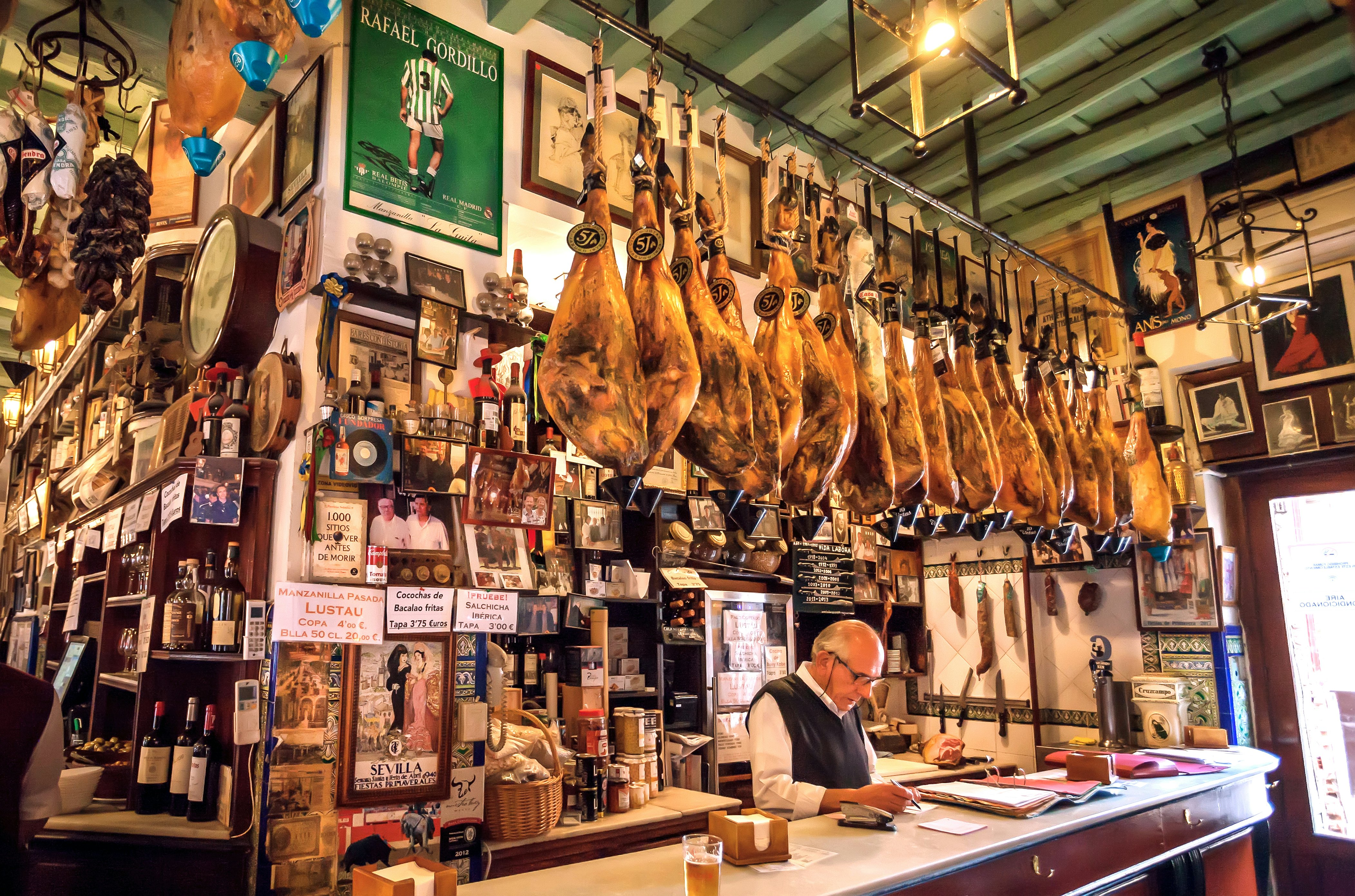A man stands behind a bar. Dried hams hang overhead, and the wall behind him is filled with photos, signs and bottles.