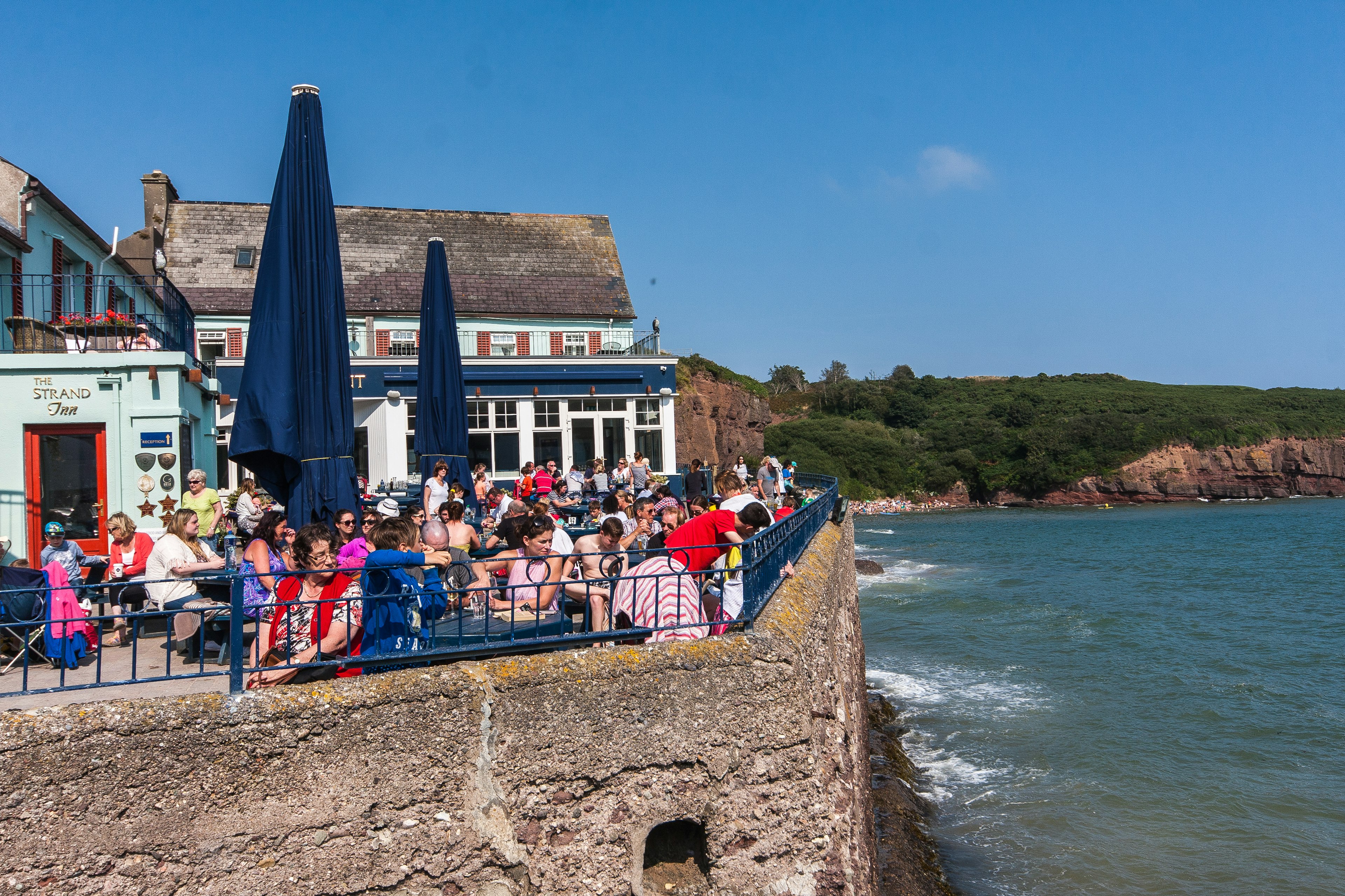 People sit on a terrace atop a seawall over the ocean. Painted houses are next to the terrace, and waves crash on the shore below.