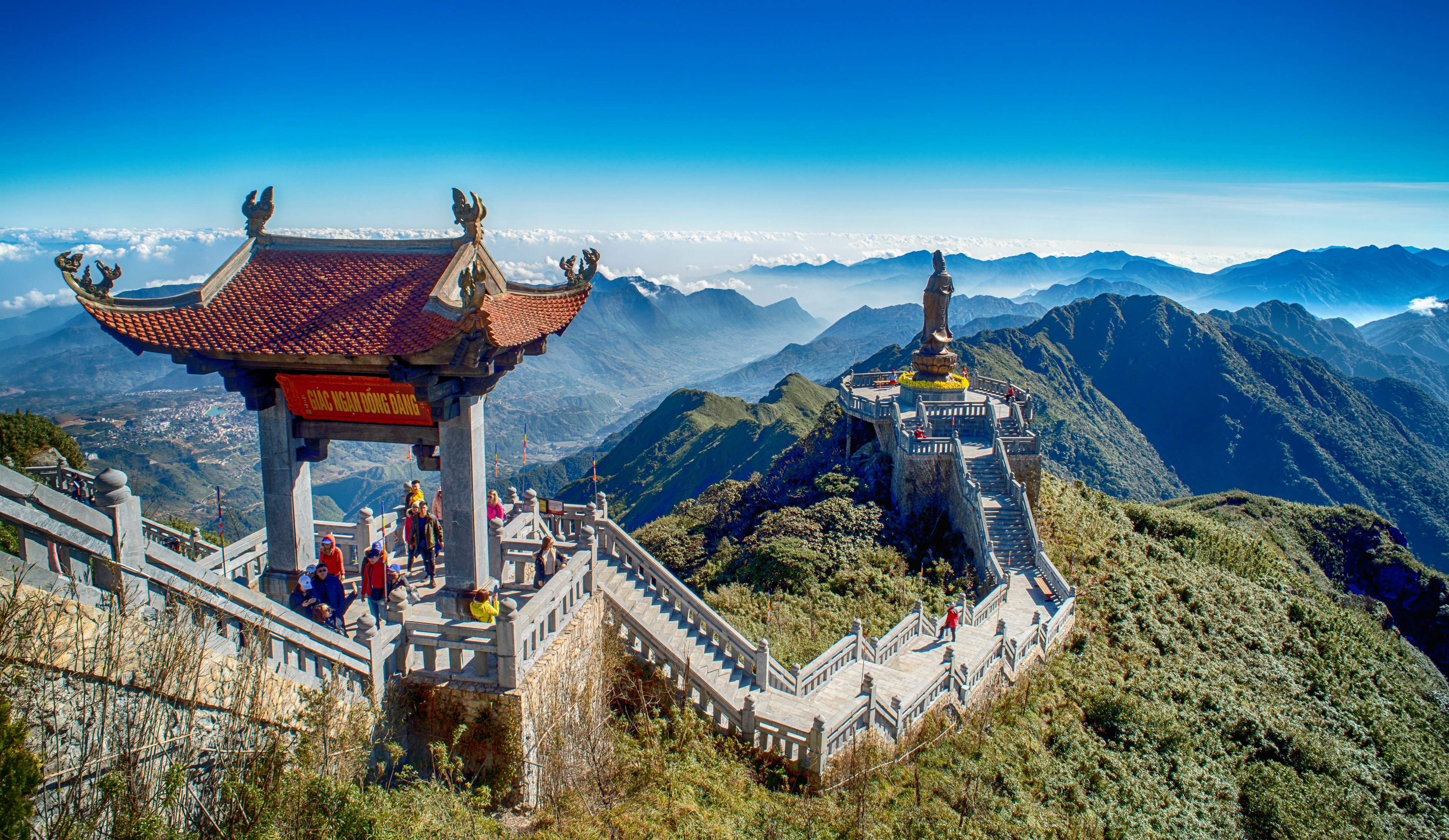 A red-roofed archway stand over a staircase leading down to the great buddha statue at mt fanSpan in Vietnam, with blue haze over the mountain range behind