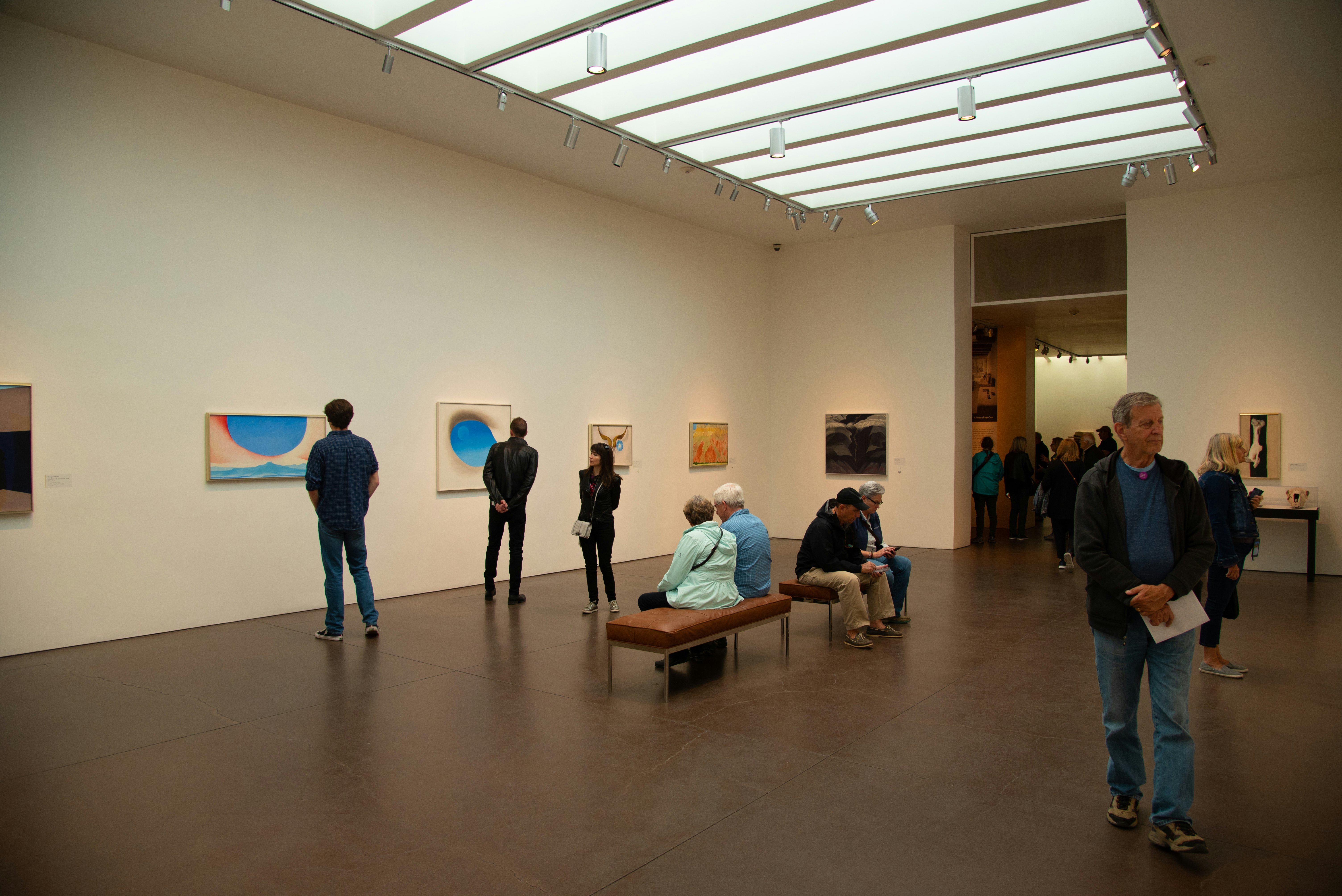 People in an art gallery sit or walk around looking at framed prints and watercolors mounted on the walls