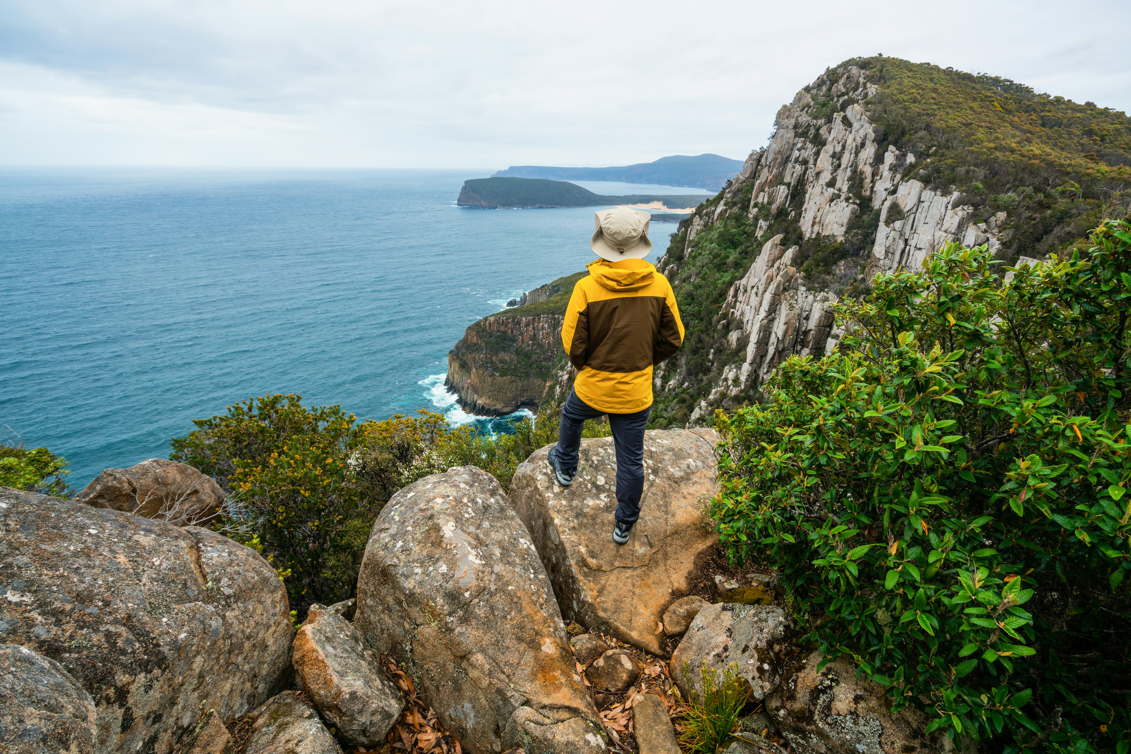 A solo hiker stands on a rock looking out of a rocky coastline