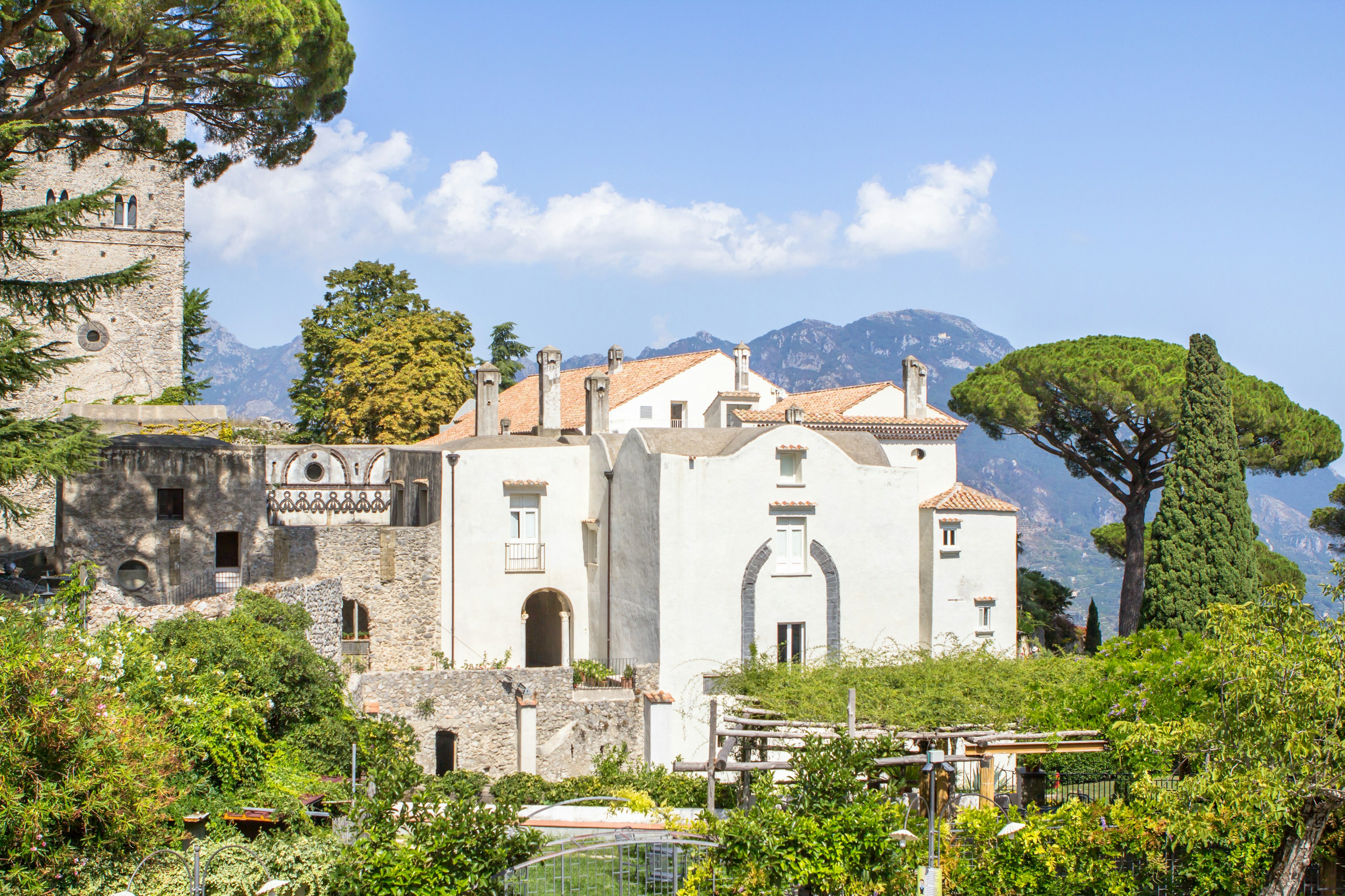 Facade of an ornate villa with trees in the foreground and mountain in the background.