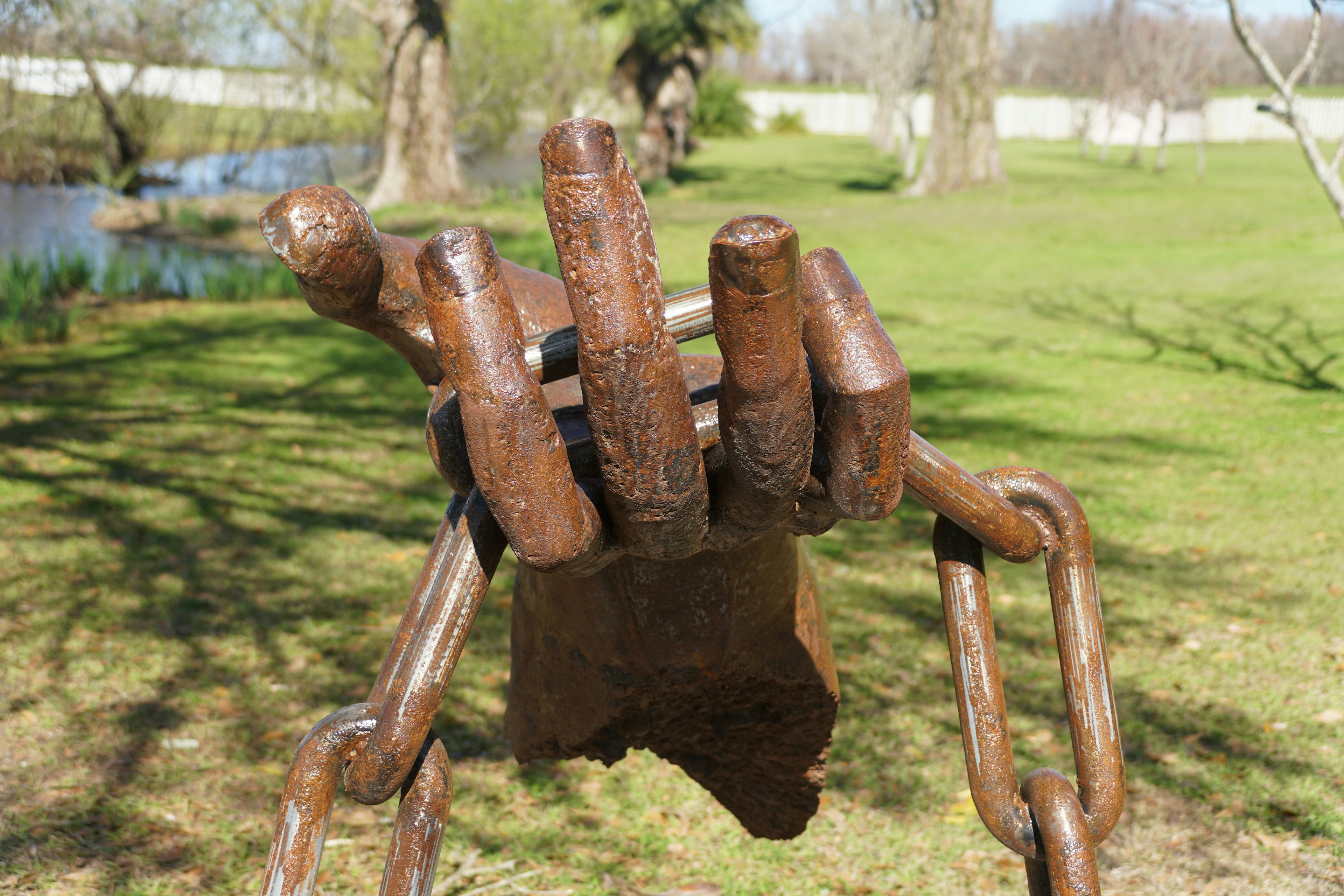 A bronze sculpture depicts a giant human hand holding chains, on a lawn by a pond