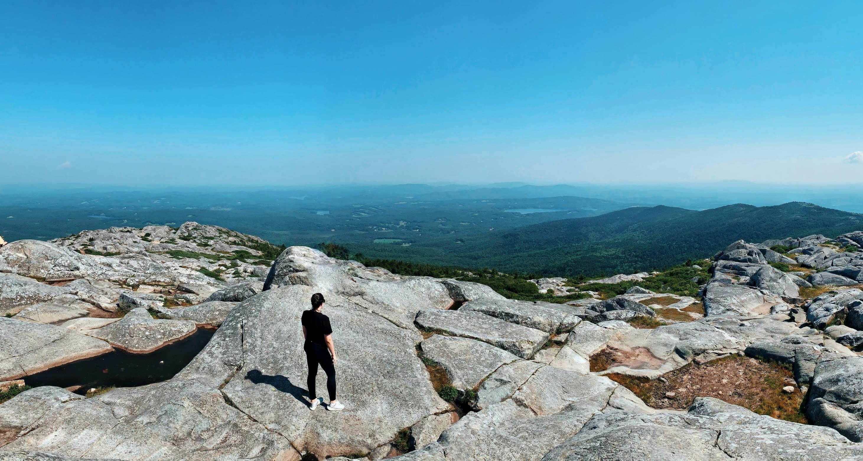 A hiker stands on the flat rocky top of a mountain and gazes out at a view that stretches for miles including hills woodland and lakes